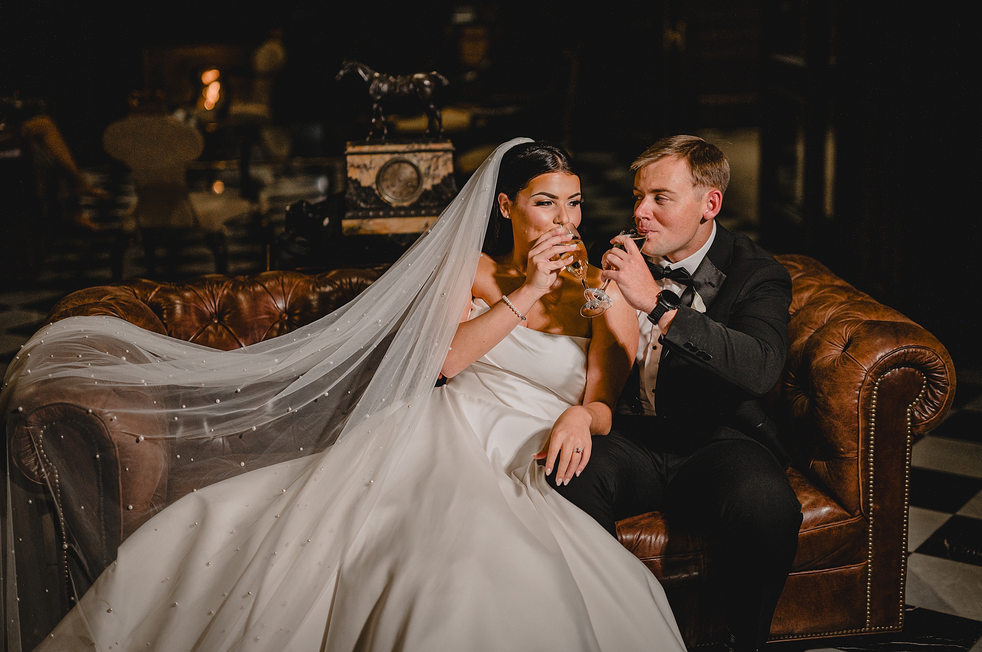 newlyweds drink glasses of wine while sitting on leather couch 