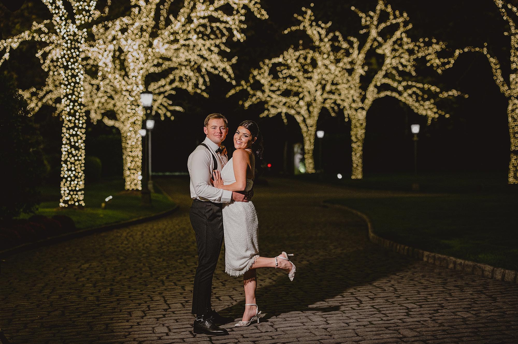 bride in short white dress hug on driveway of trees wrapped in holiday lights