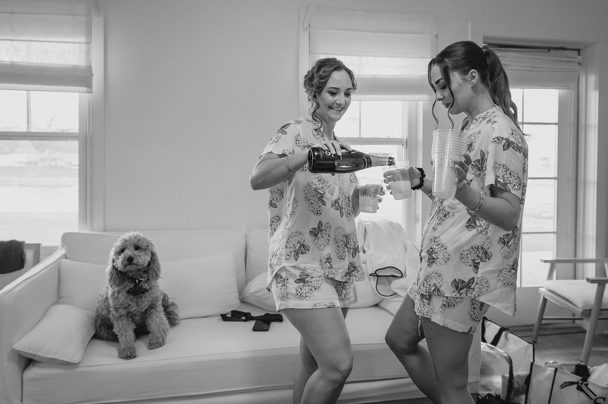 bridesmaids pour champagne while dog sits on sofa behind them 