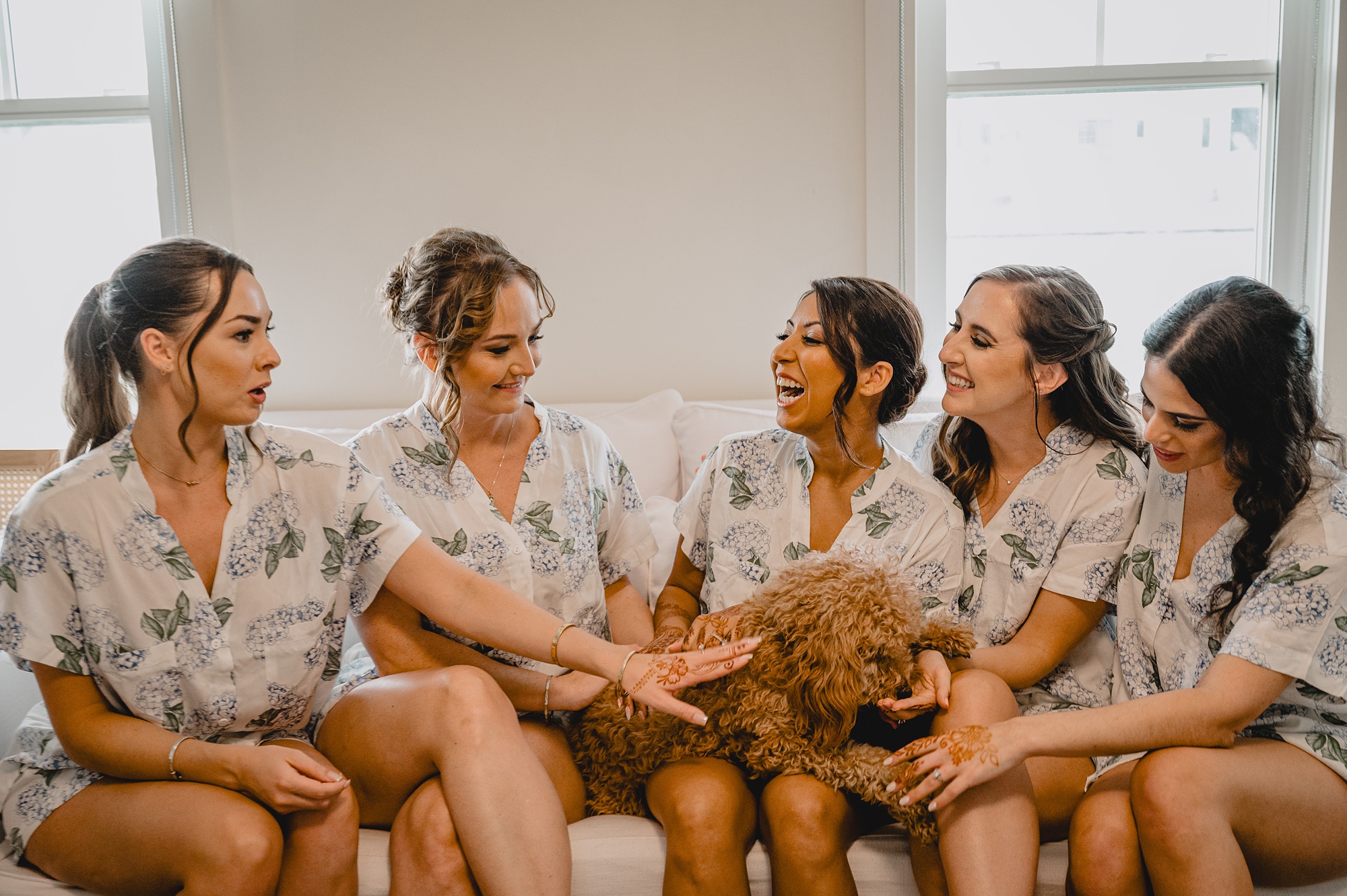 bridesmaids in floral pajamas sit with bride and her dog on sofa 