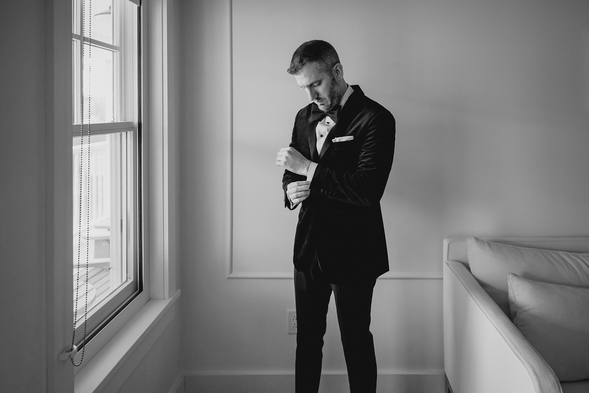 groom in black suit adjusts cufflinks standing by window at the Lake House at Canandaigua