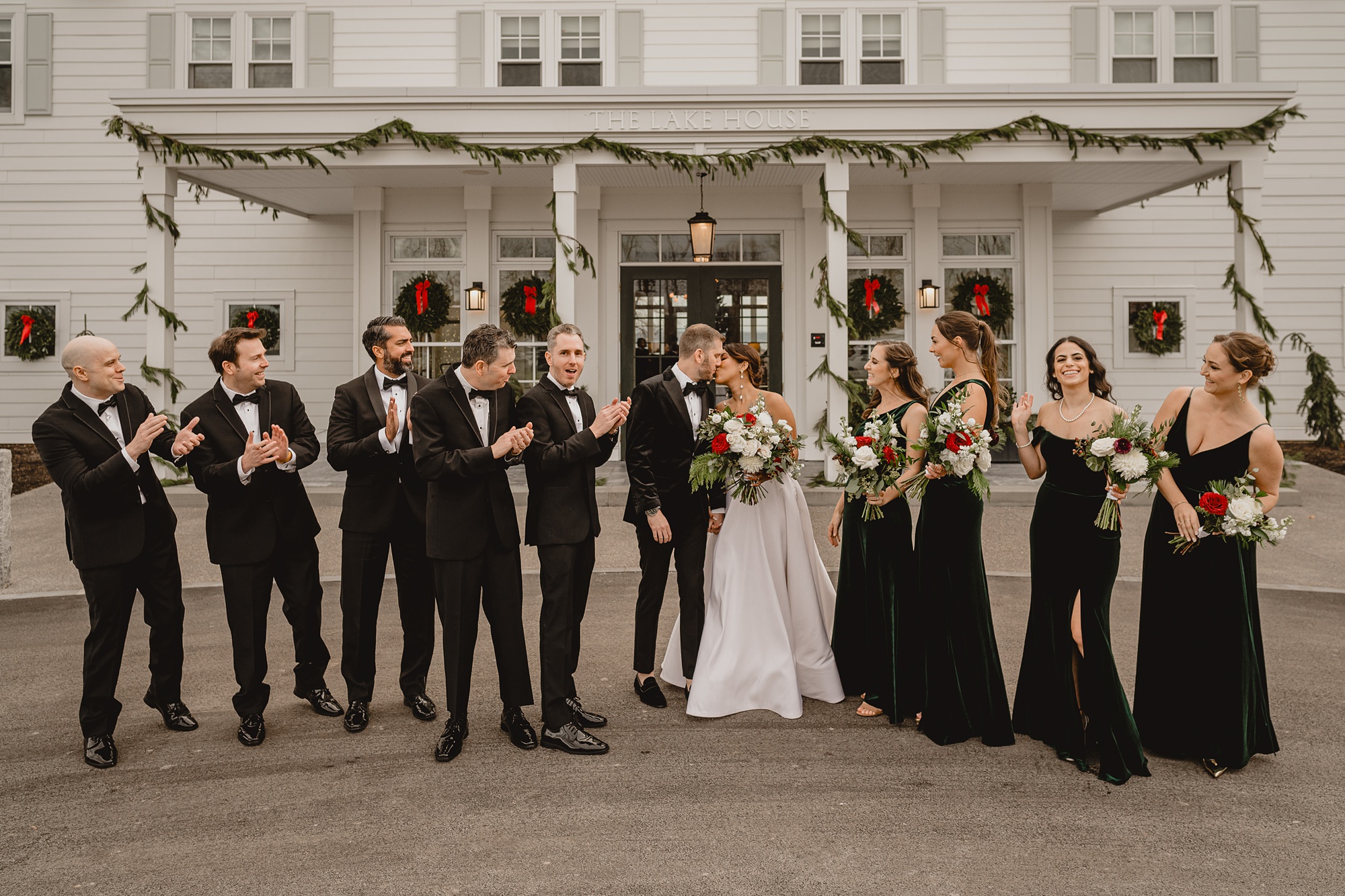 bride and groom kiss while wedding party claps around them in front of the Lake House at Canandaigua