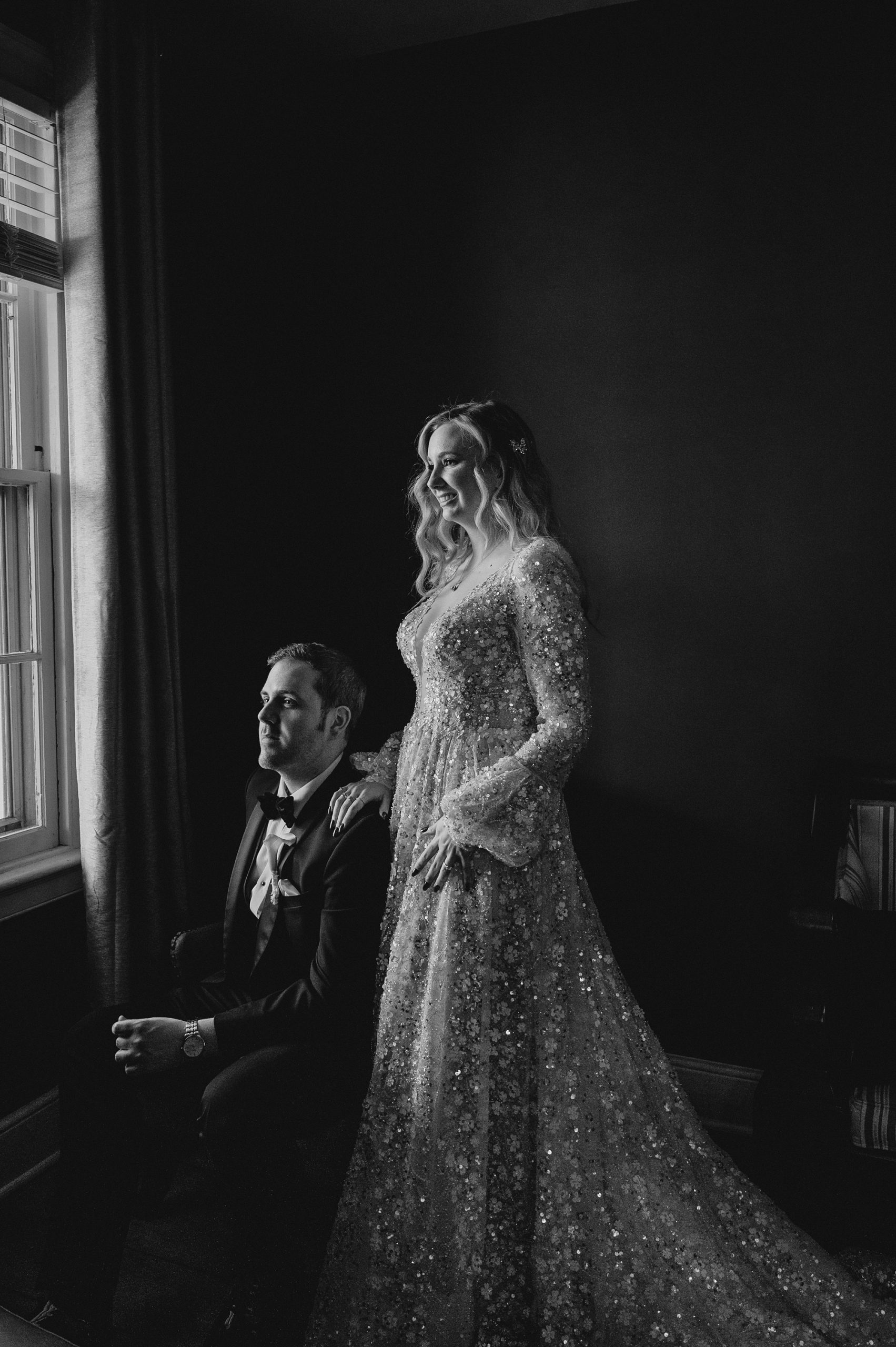 bride stands behind groom looking out window in black and white photo