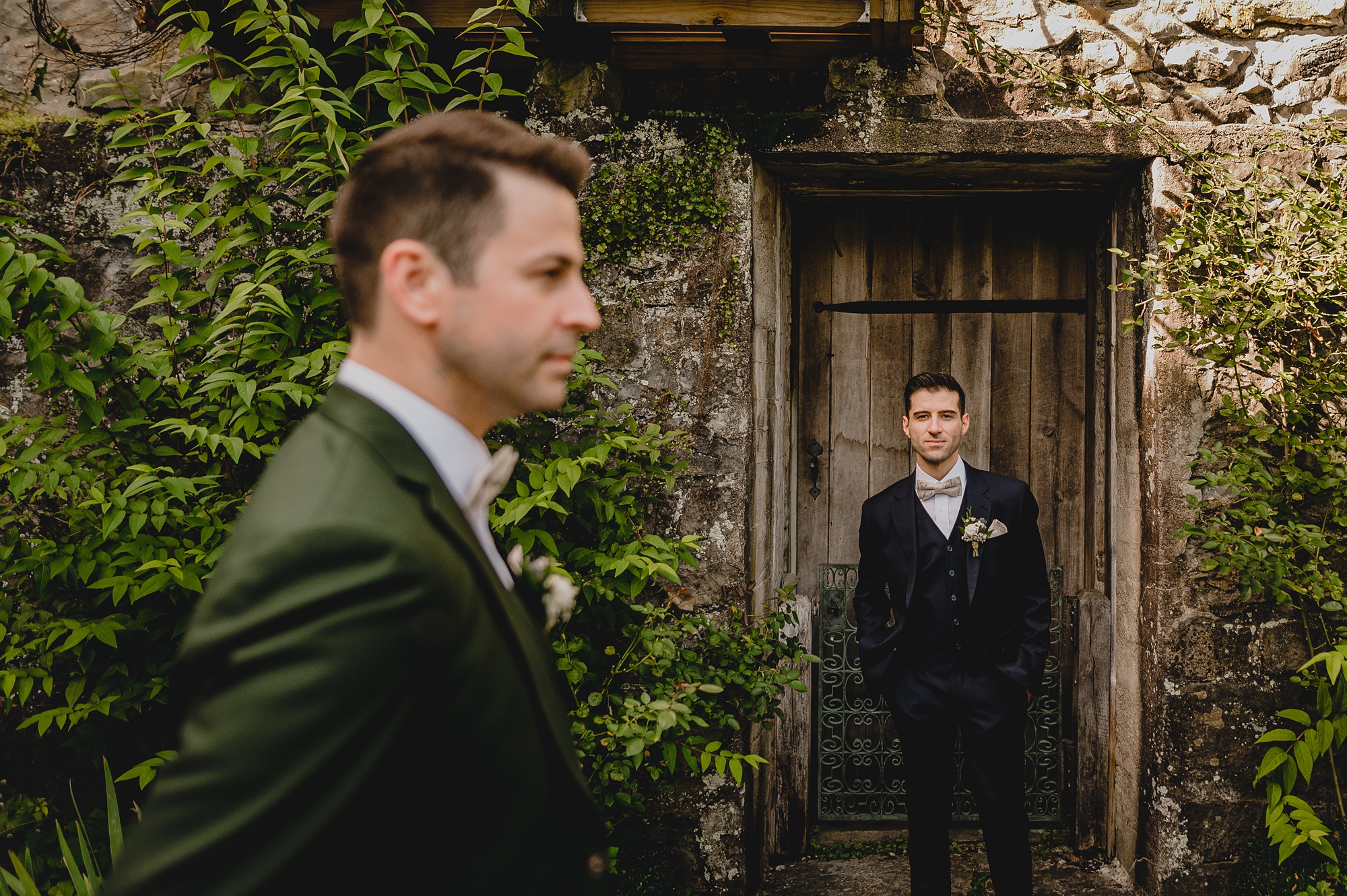 groom leans against wooden door while husband looks across building