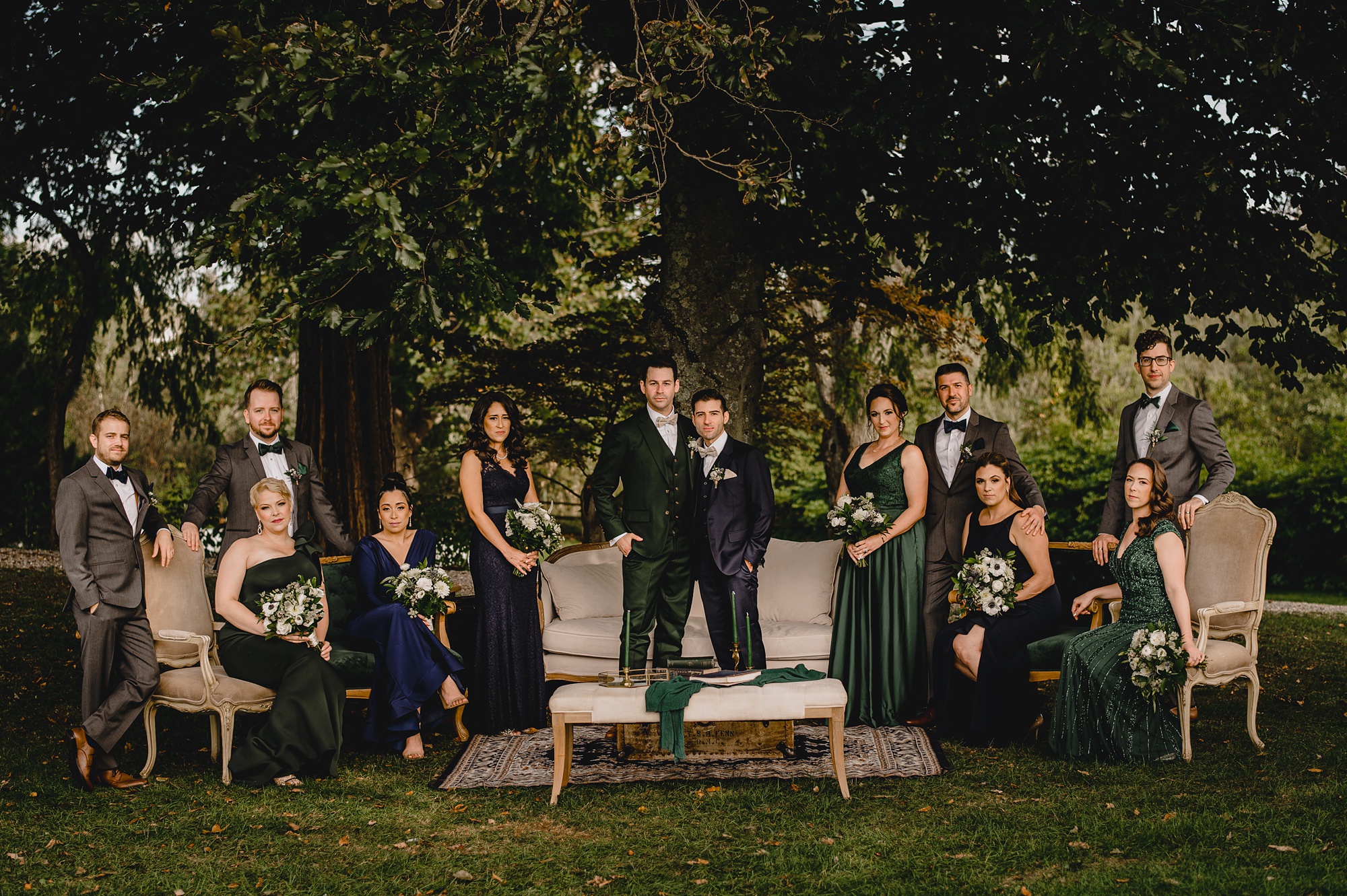 grooms pose with wedding party standing near ivory couches and seating under trees at Crossed Keys Estate