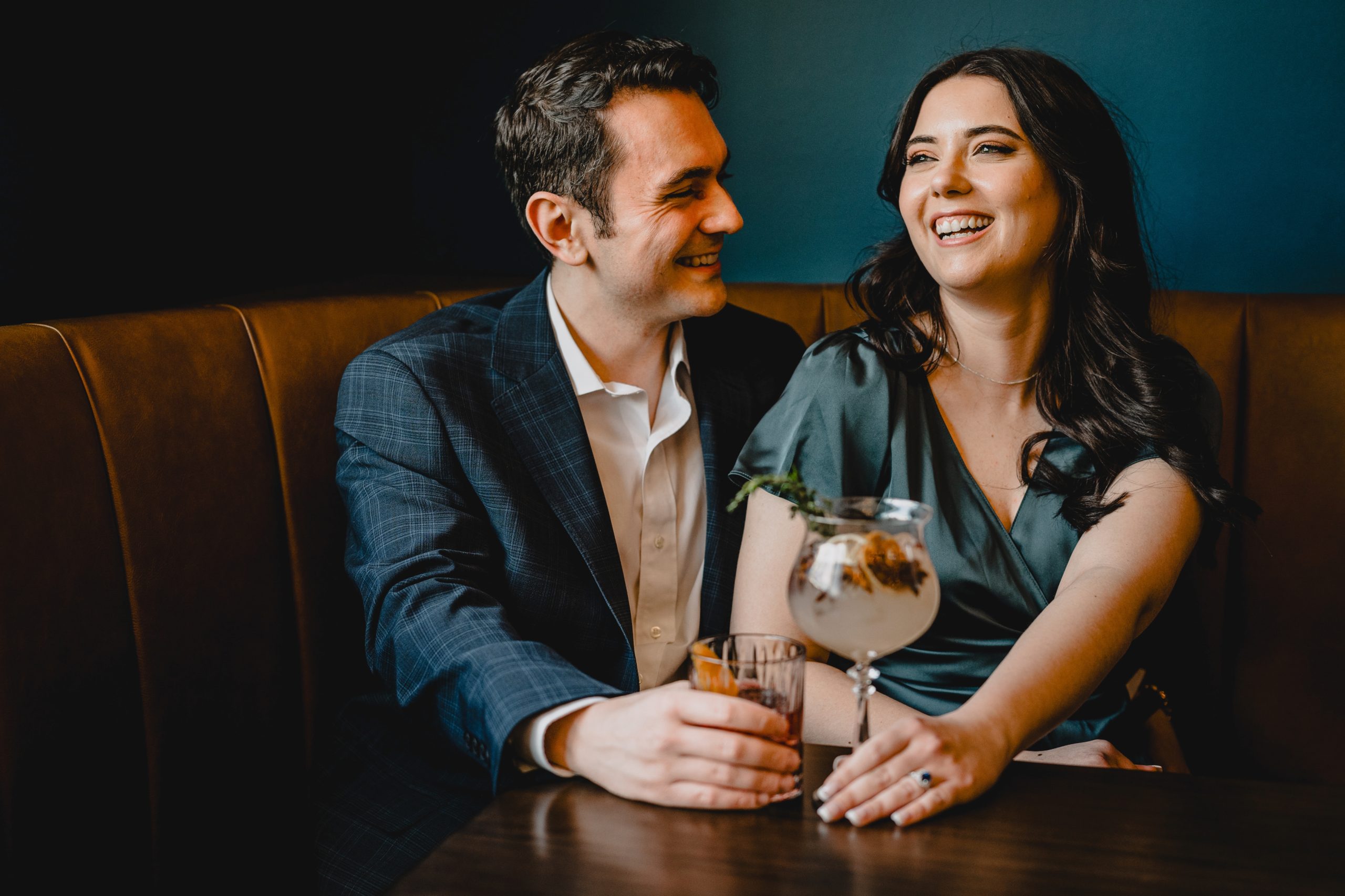 man and woman laugh sitting in NYC bar with drinks during summer engagement photos 