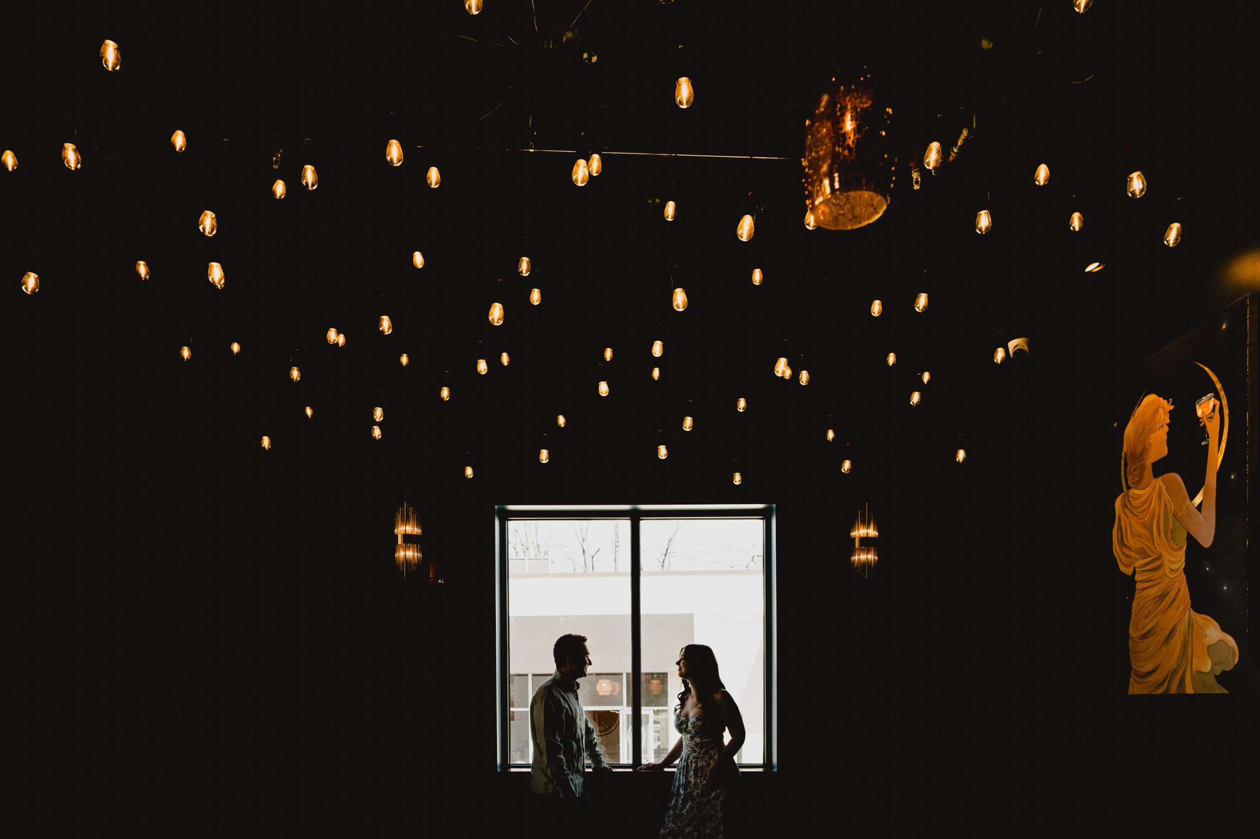 engaged couple poses in window of NYC bar with lights hanging overhead