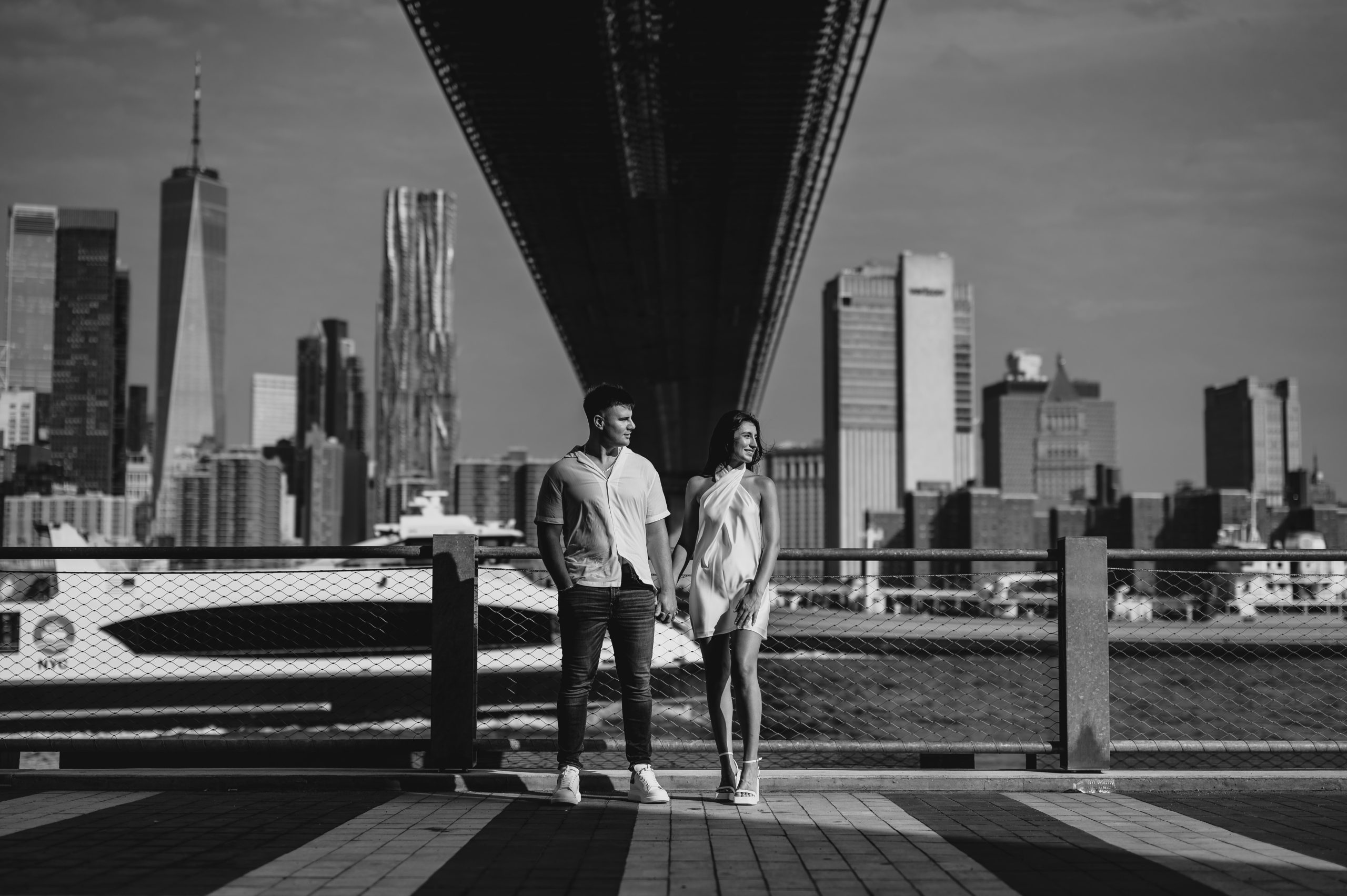 engaged couple holds hands under Brooklyn Bridge in front of NYC skyline