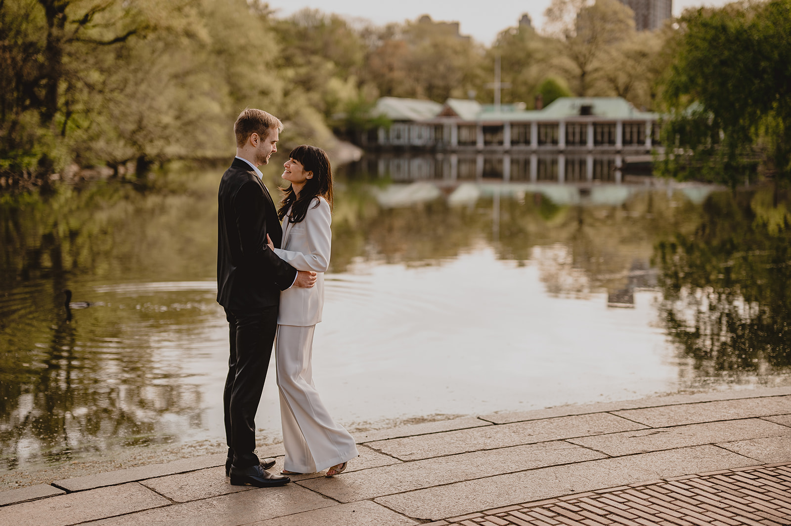 engaged couple stands by pond near Boat House in Central Park