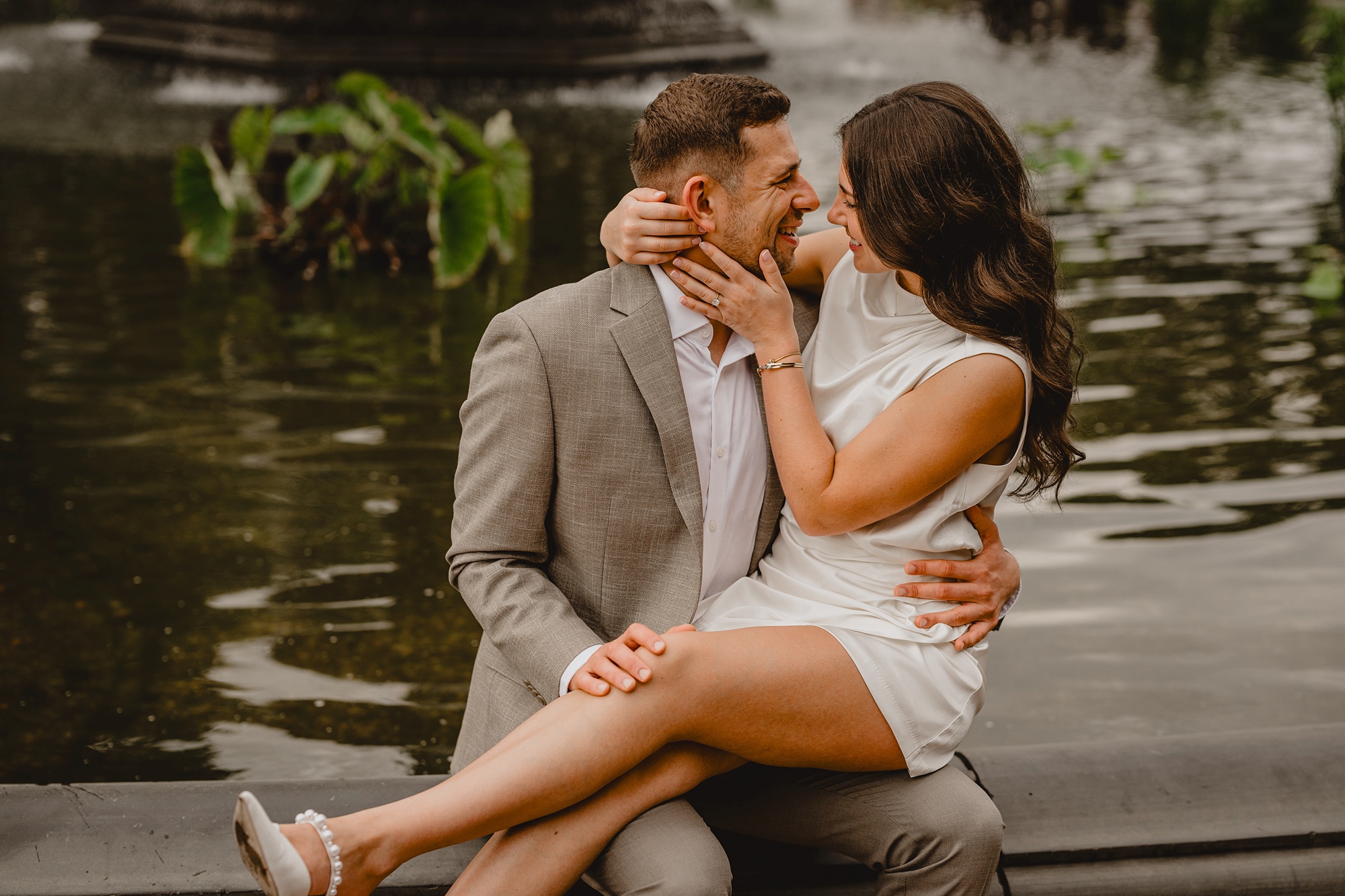 woman in ivory dress sits on man's lap on edge of Bethesda fountain in Central Park 