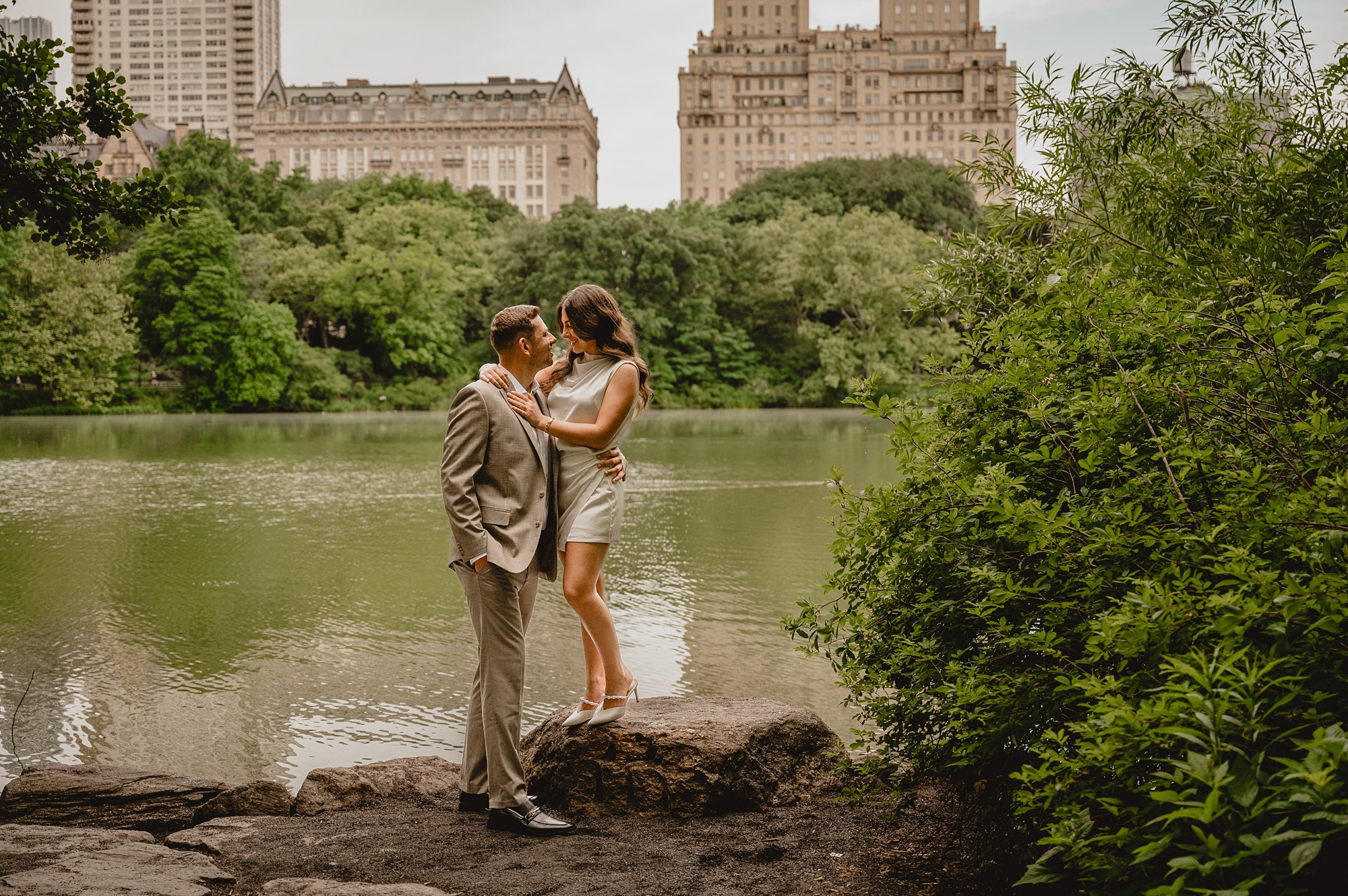 woman stands on rock hugging man in front of pond in Central Park