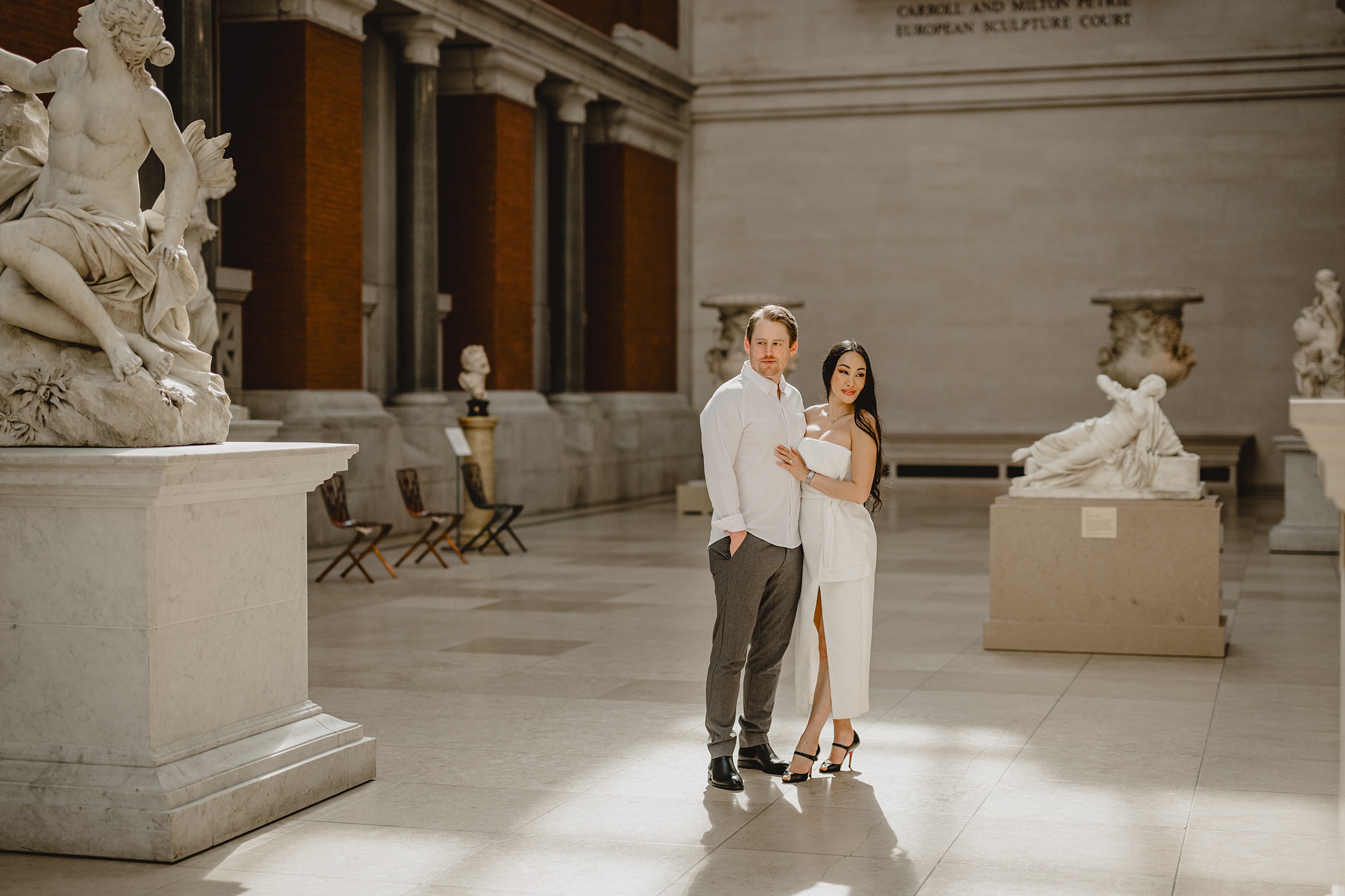 engaged couple stands between statues at the Met in NYC