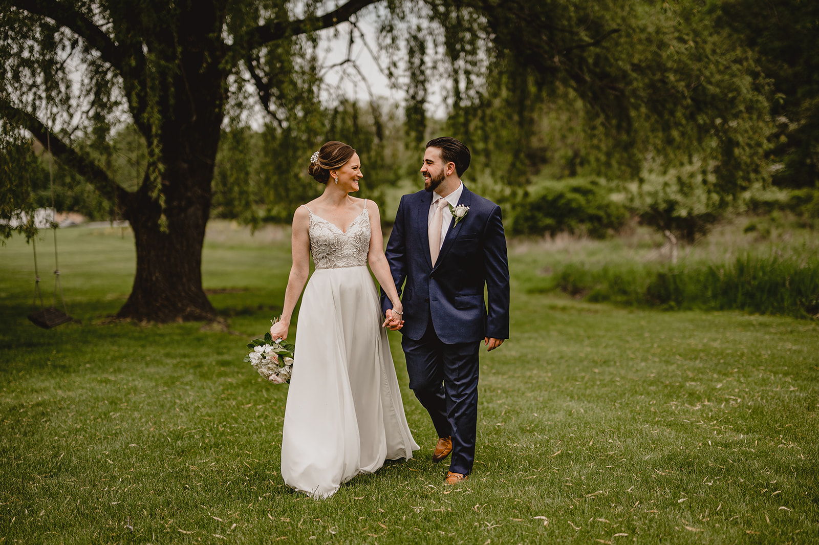 couple holds hands walking across lawn under weeping willow tree