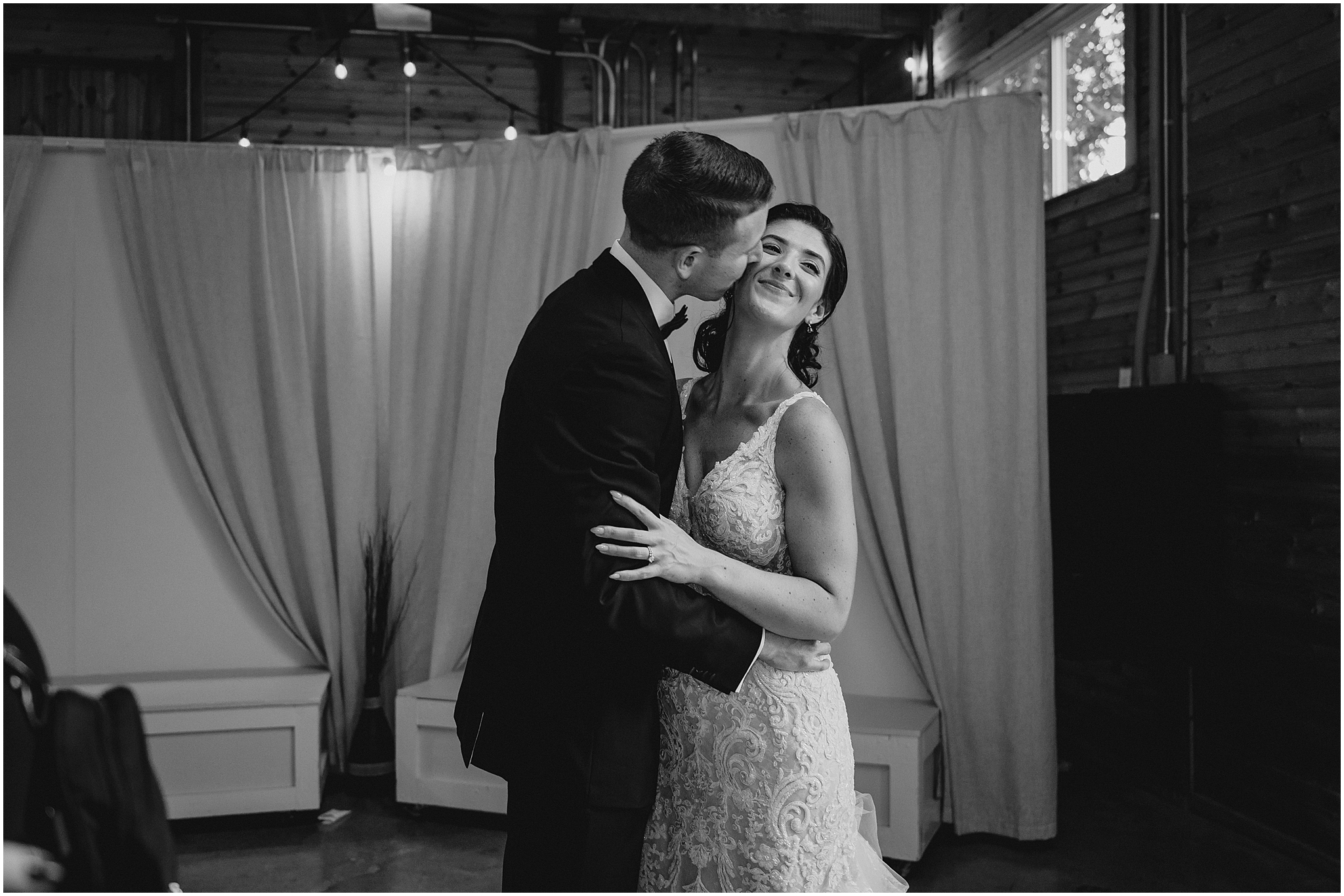 groom kisses bride's cheek in front of hanging white fabric at reception