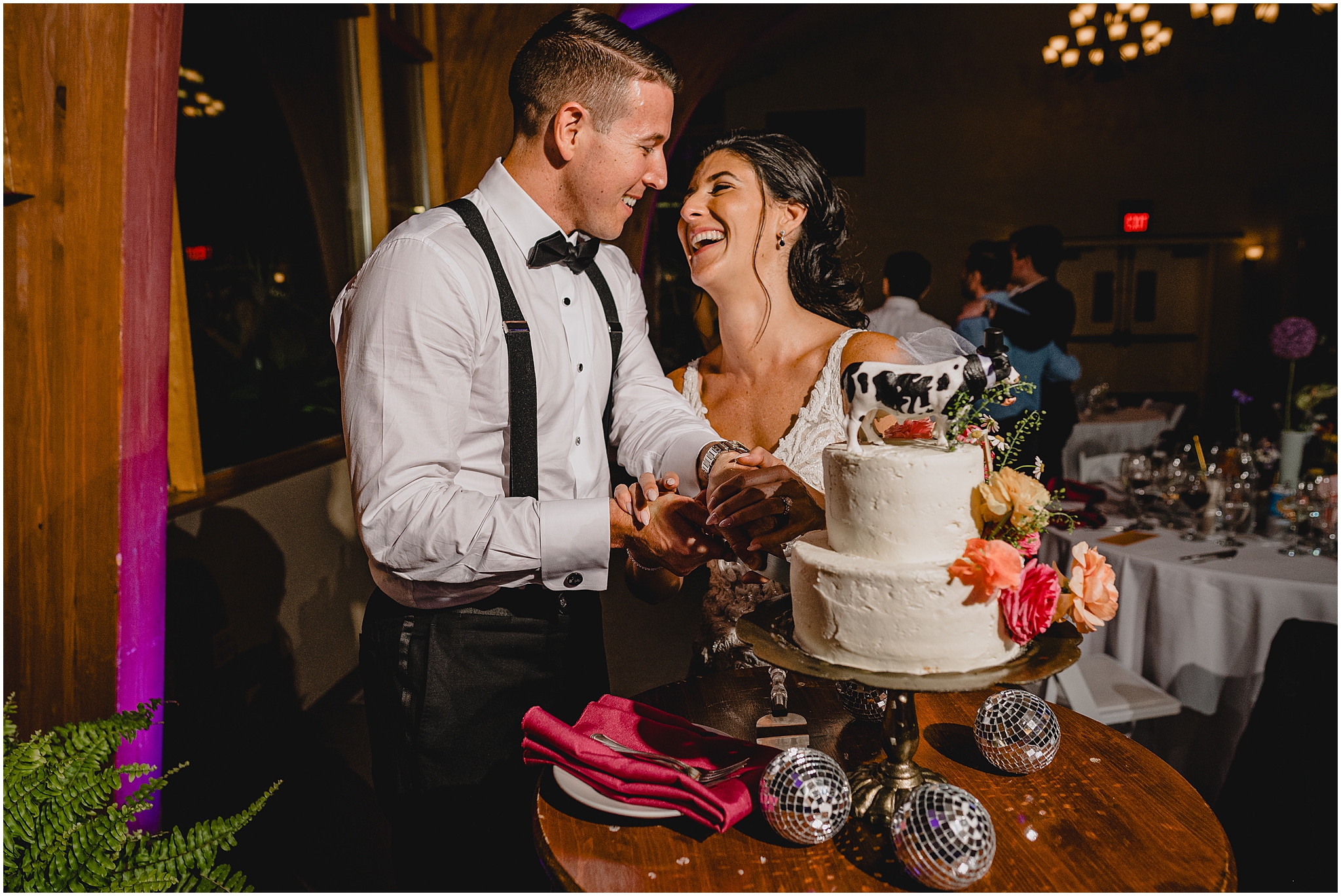newlyweds laugh during wedding reception and cutting cake with cow cake topper