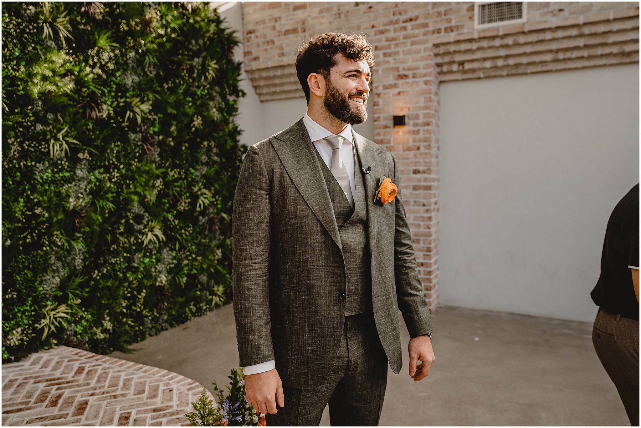 groom in grey suit with coral rose boutonnière stands outside the Perona Farms Refinery 