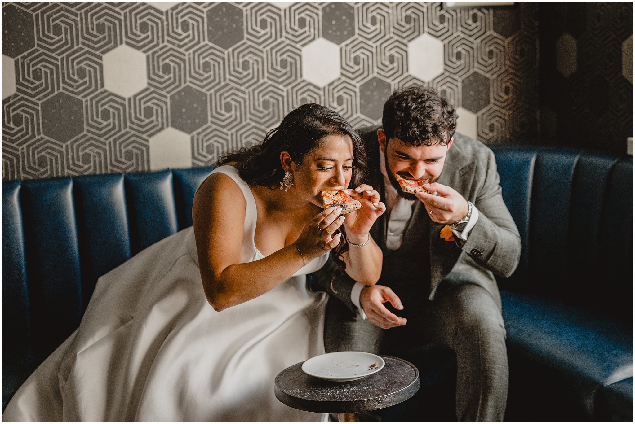 newlyweds eat slices of pizza together during wedding day on blue leather couch
