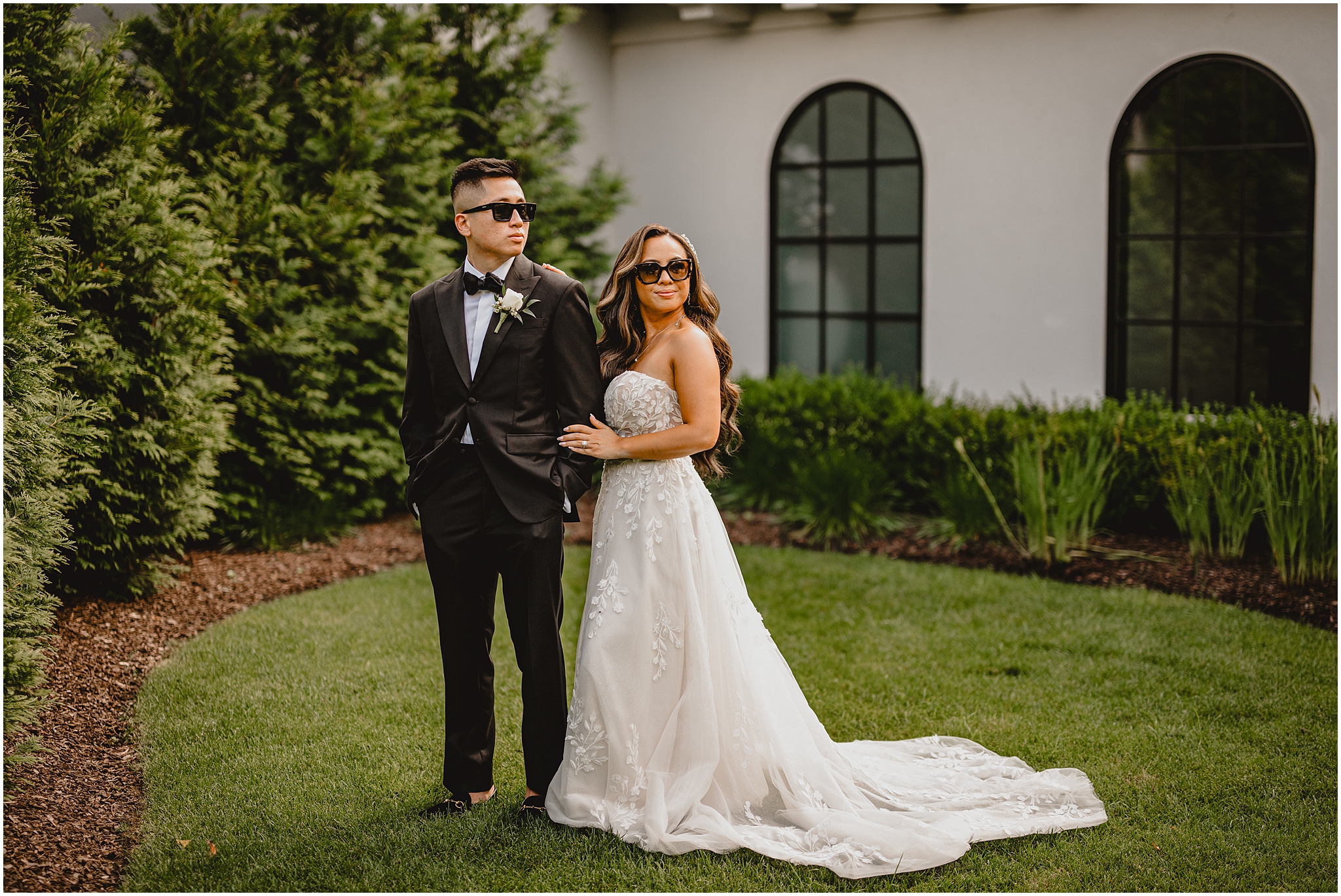 newlyweds wearing sunglasses pose in courtyard in front of white walls at the Refinery at Perona Farms