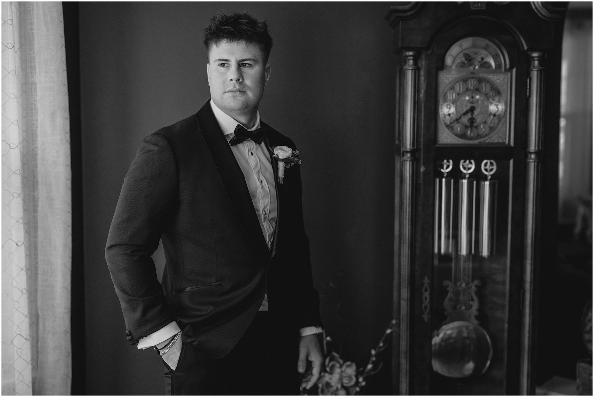 groom stands in black tux by grandfather clock at home