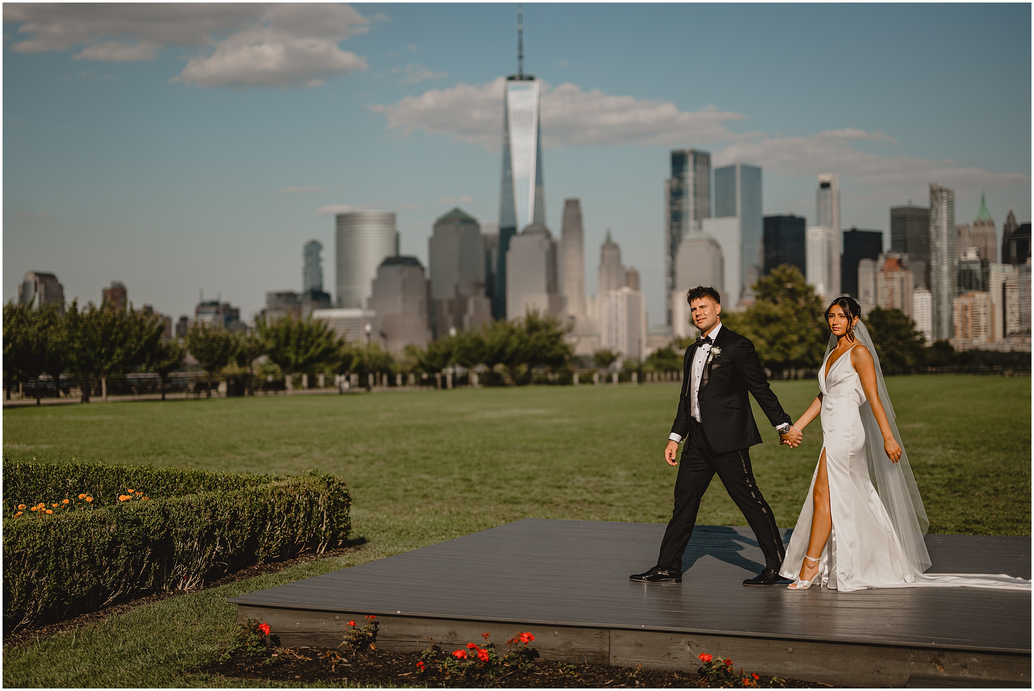 groom leads bride across wooden platform in front of Manhattan skyline in NJ