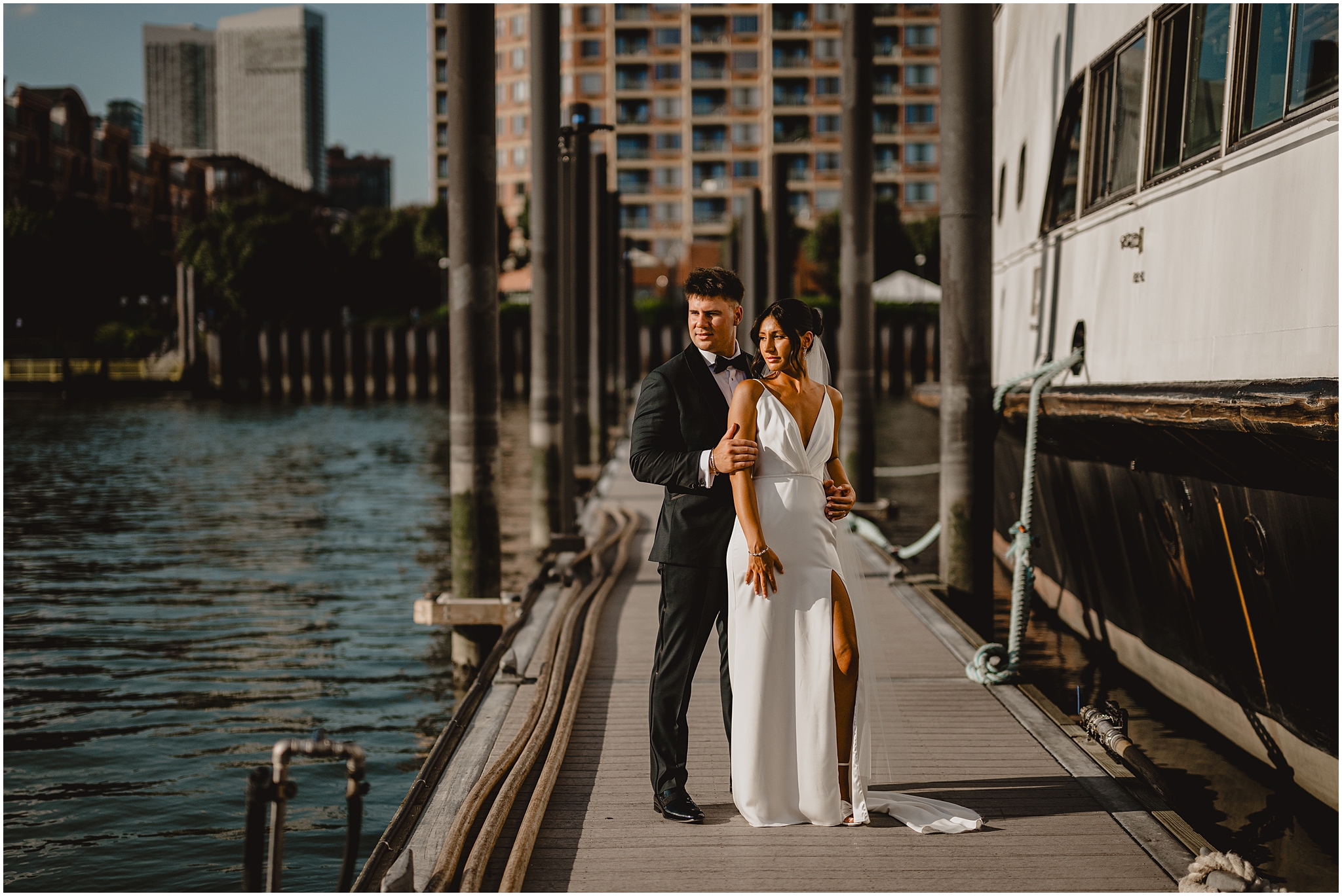 groom hugs bride from behind standing on dock near ship in Jersey City