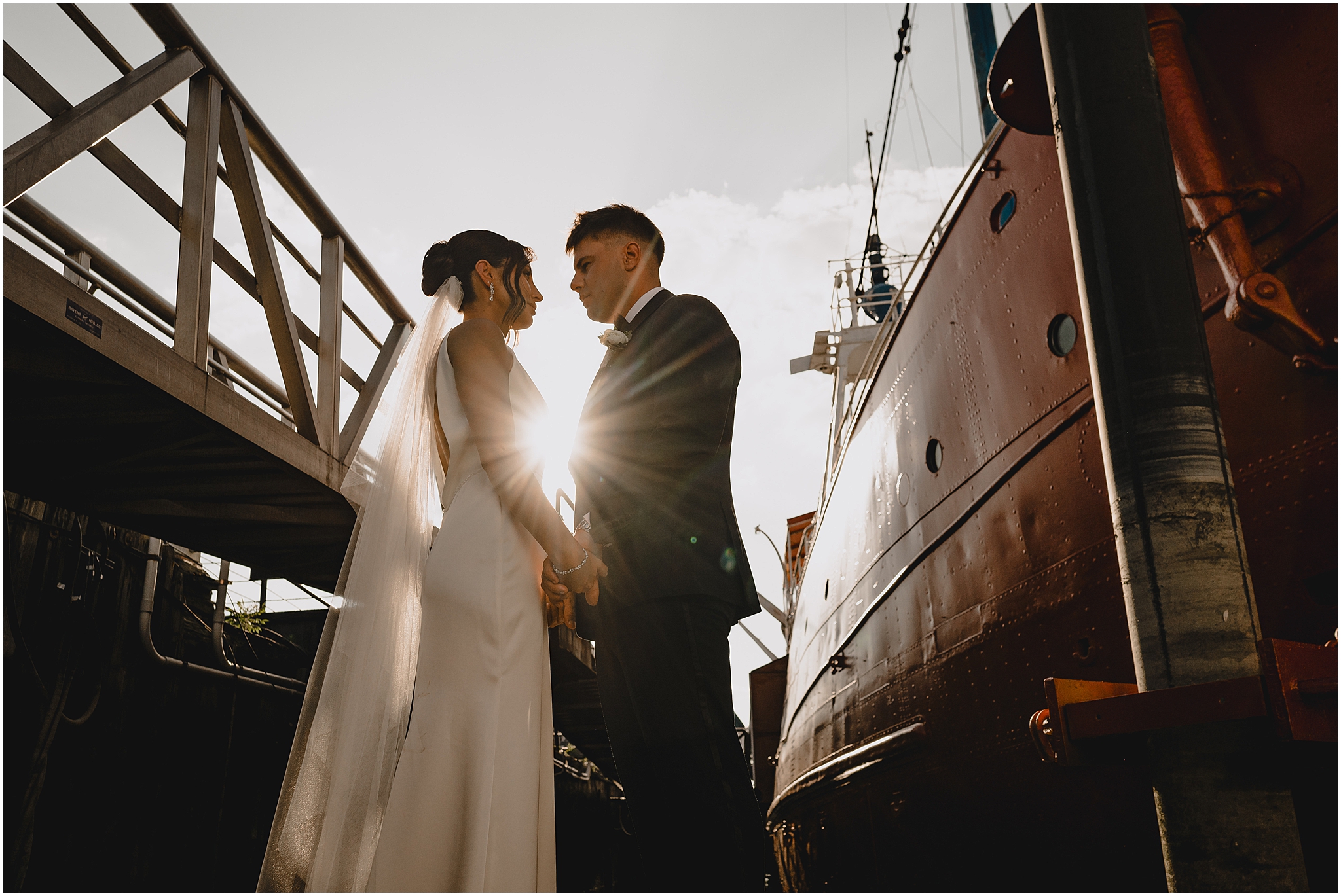 bride and groom look at each other standing by ship with sun flare between them