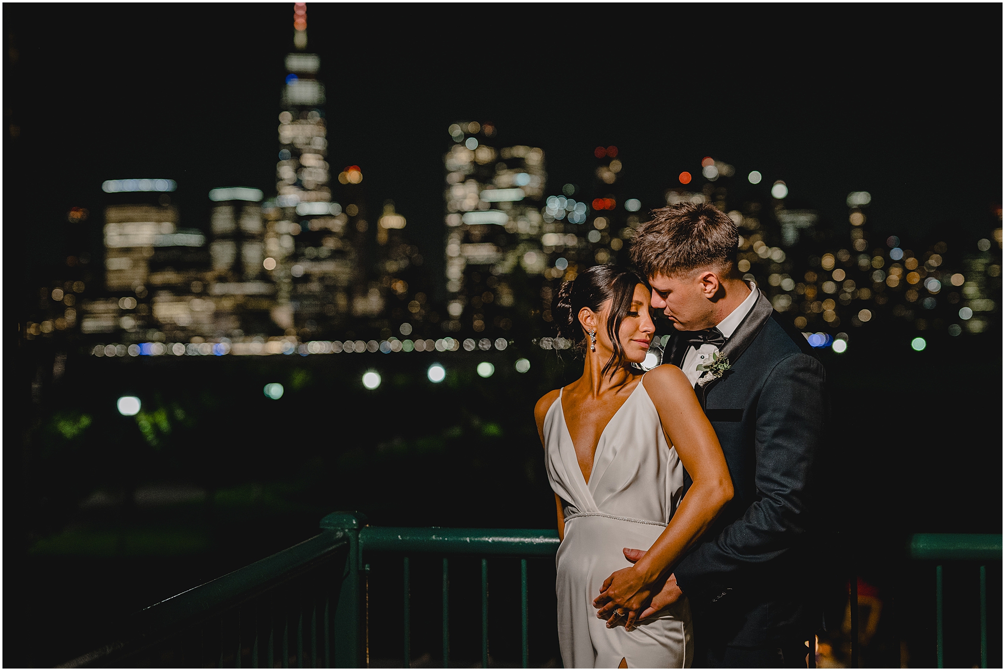 groom hugs bride from behind on balcony in front of Manhattan skyline at night