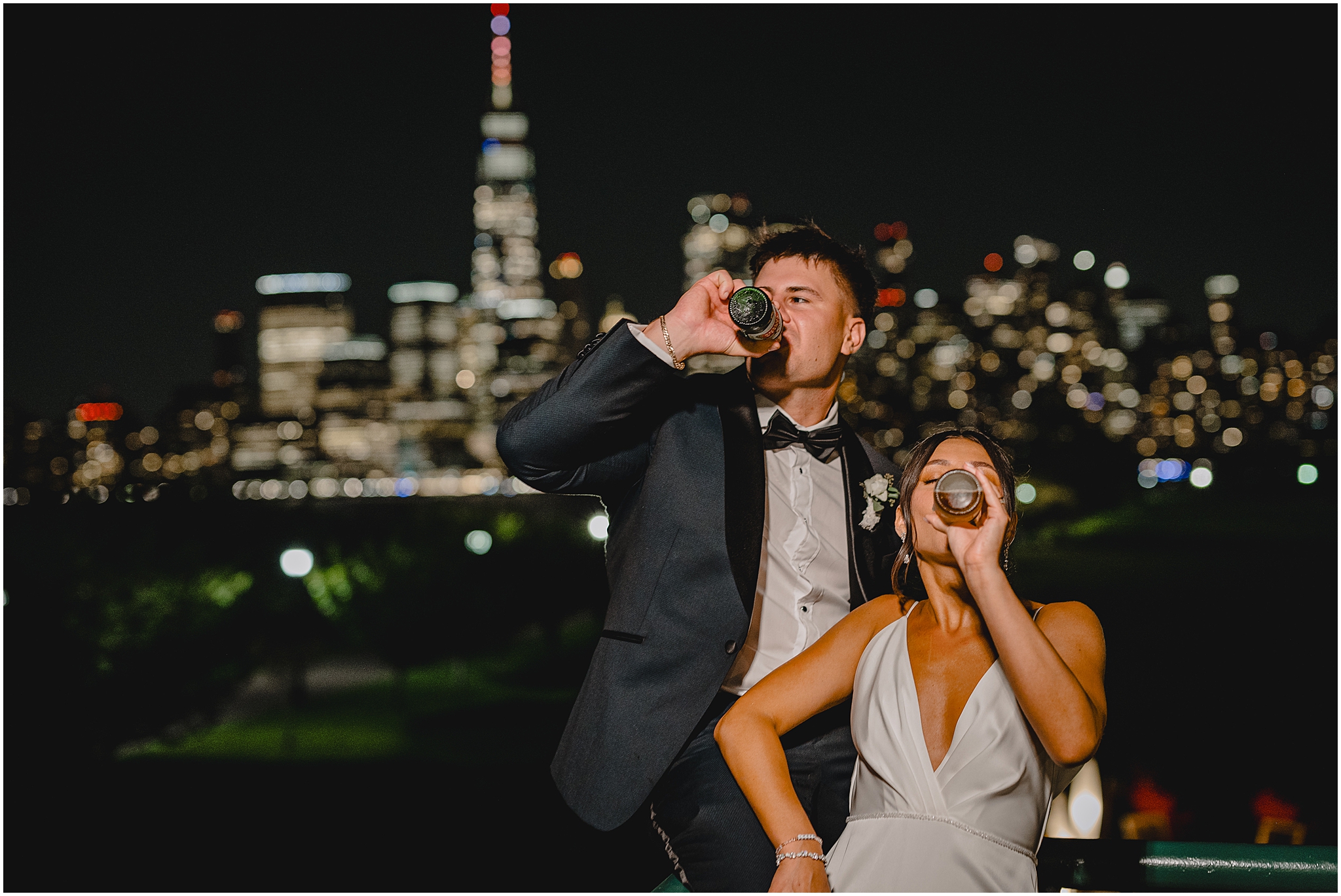 bride and groom drink beer standing on balcony in front of NYC skyline at night