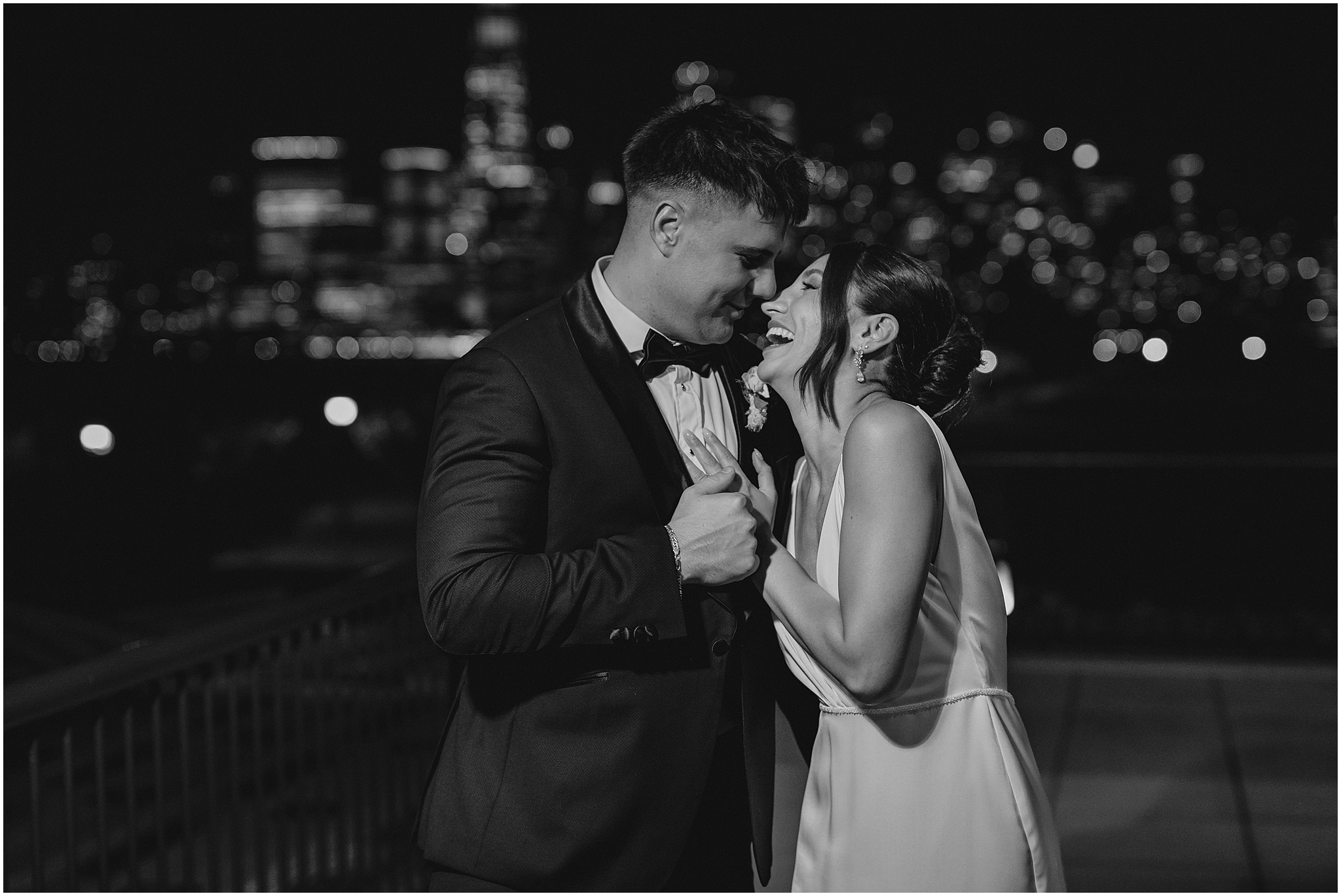 bride and groom laugh together hugging on balcony at night in front of NYC skyline 