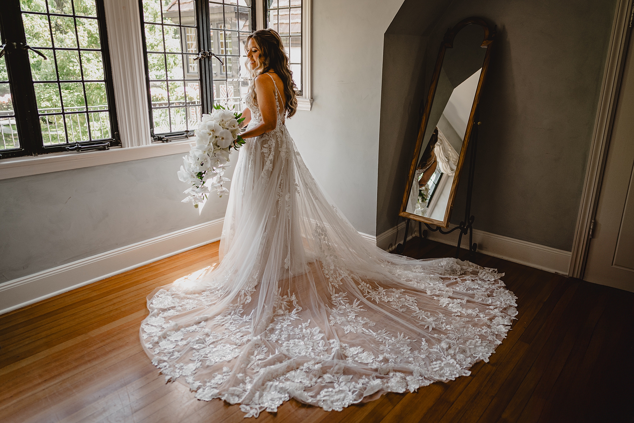 bride stands in window of suite at Pleasantdale Chateau holding casting bouquet of orchids