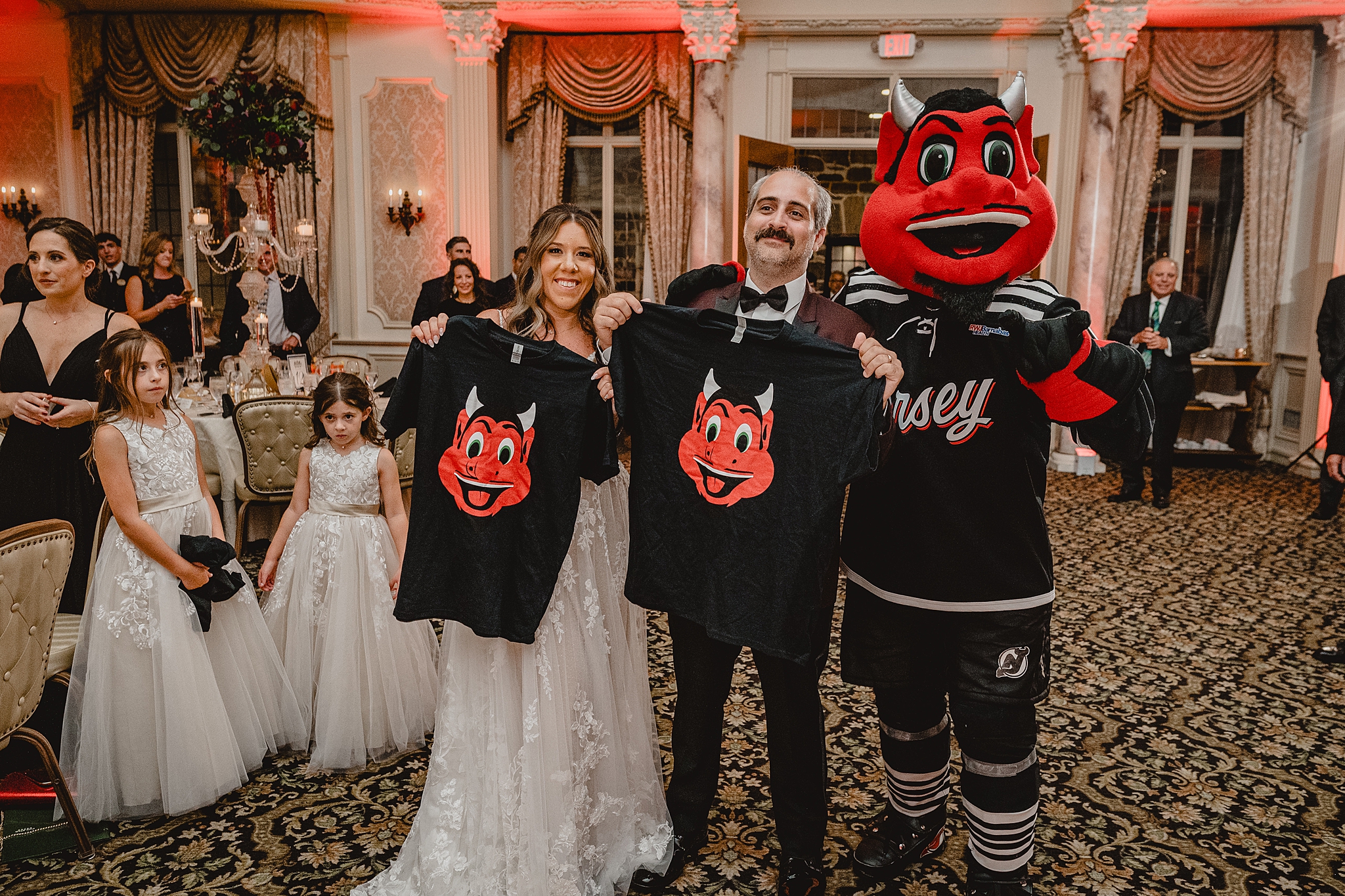 bride and groom hold up Red Devils shirts and pose with mascot in ballroom at Pleasantdale Chateau