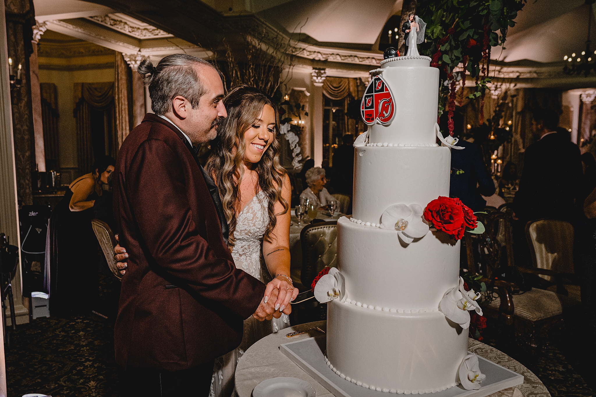 newlyweds cut tiered wedding cake with white and rd flowers and Red Devil mascot on top 