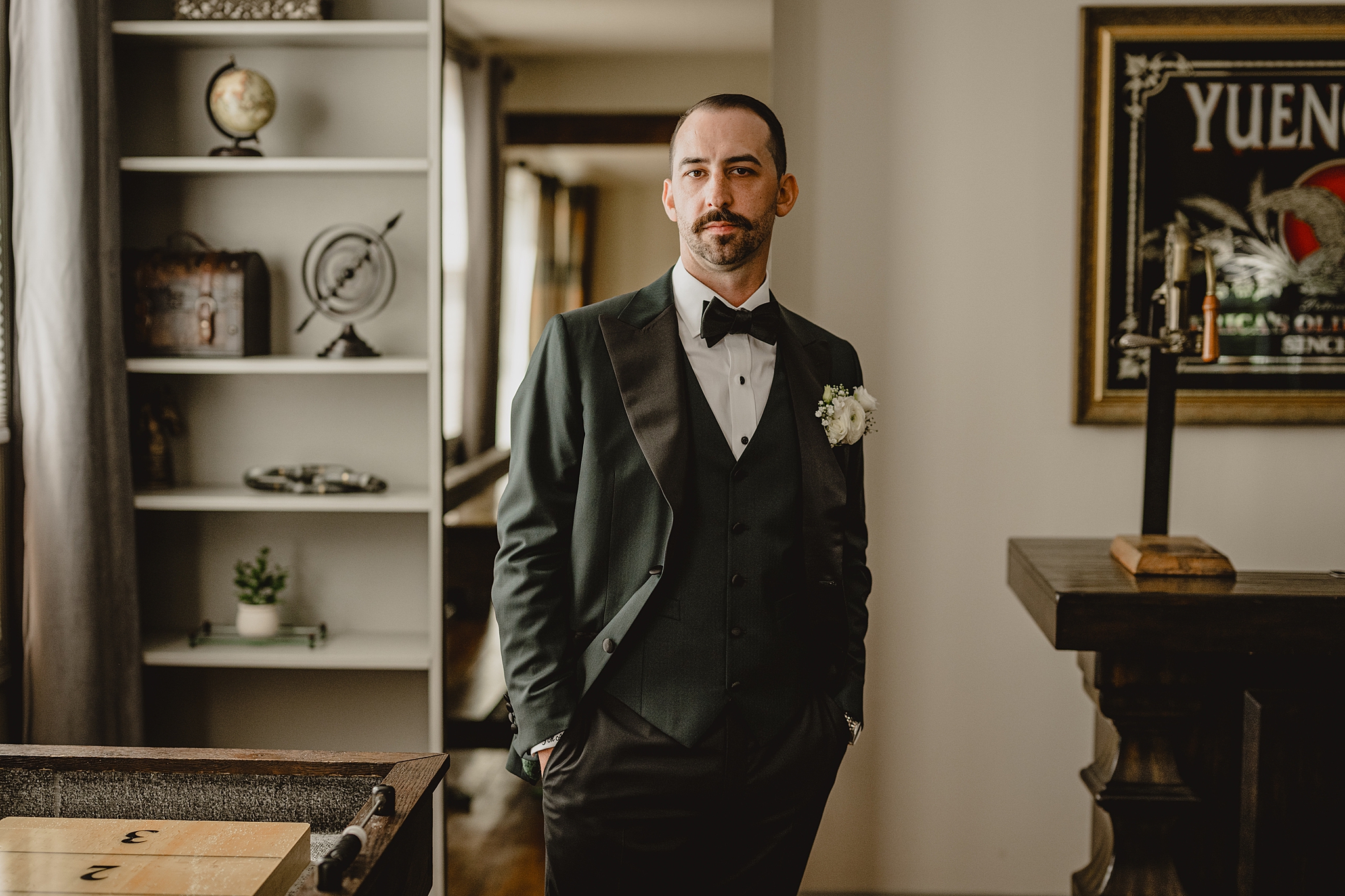 groom stands with hands in dark green suit in groom's suite at David's Country Inn