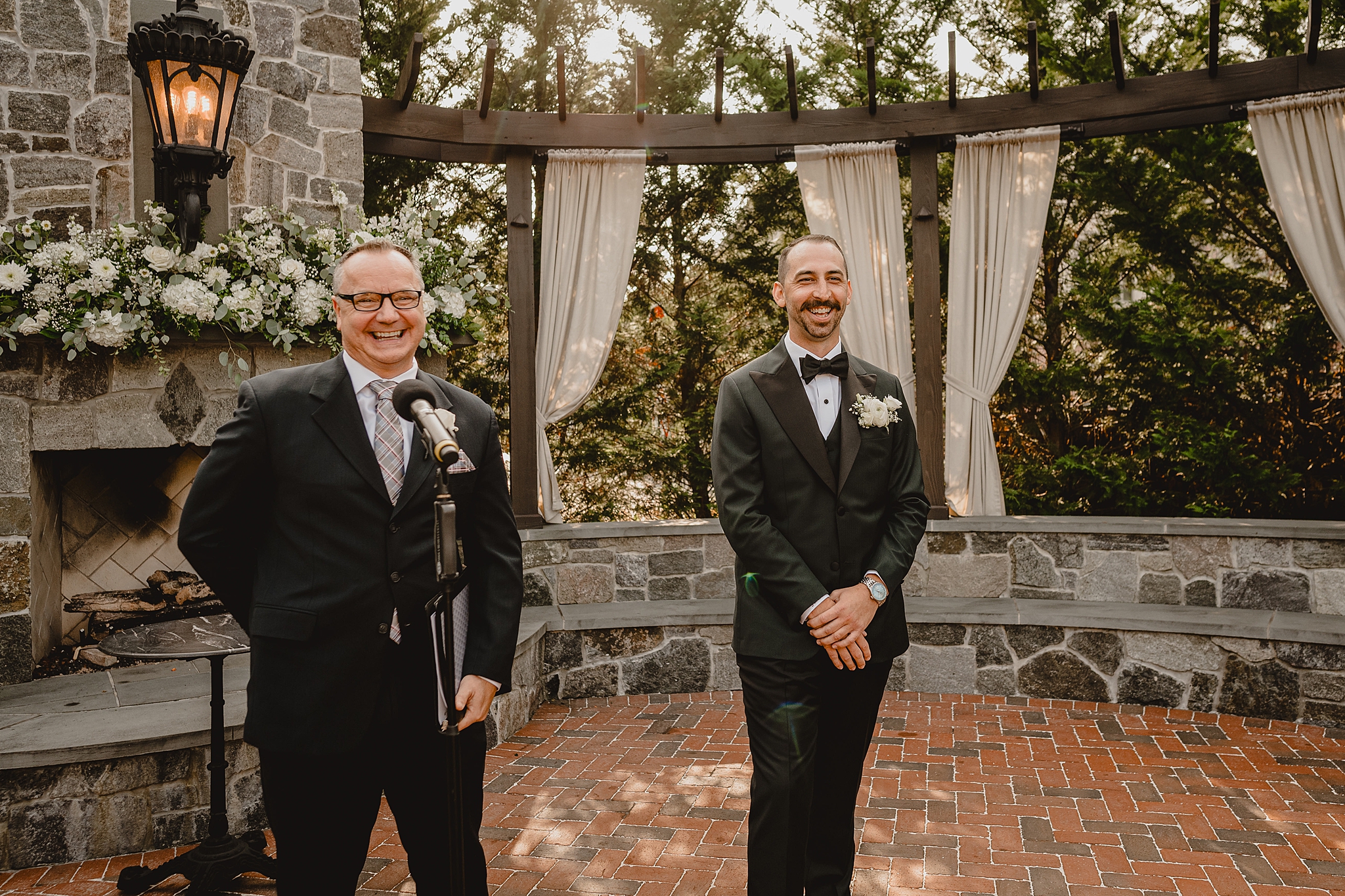 groom laughs with officiant waiting for ceremony on brick patio by fireplace