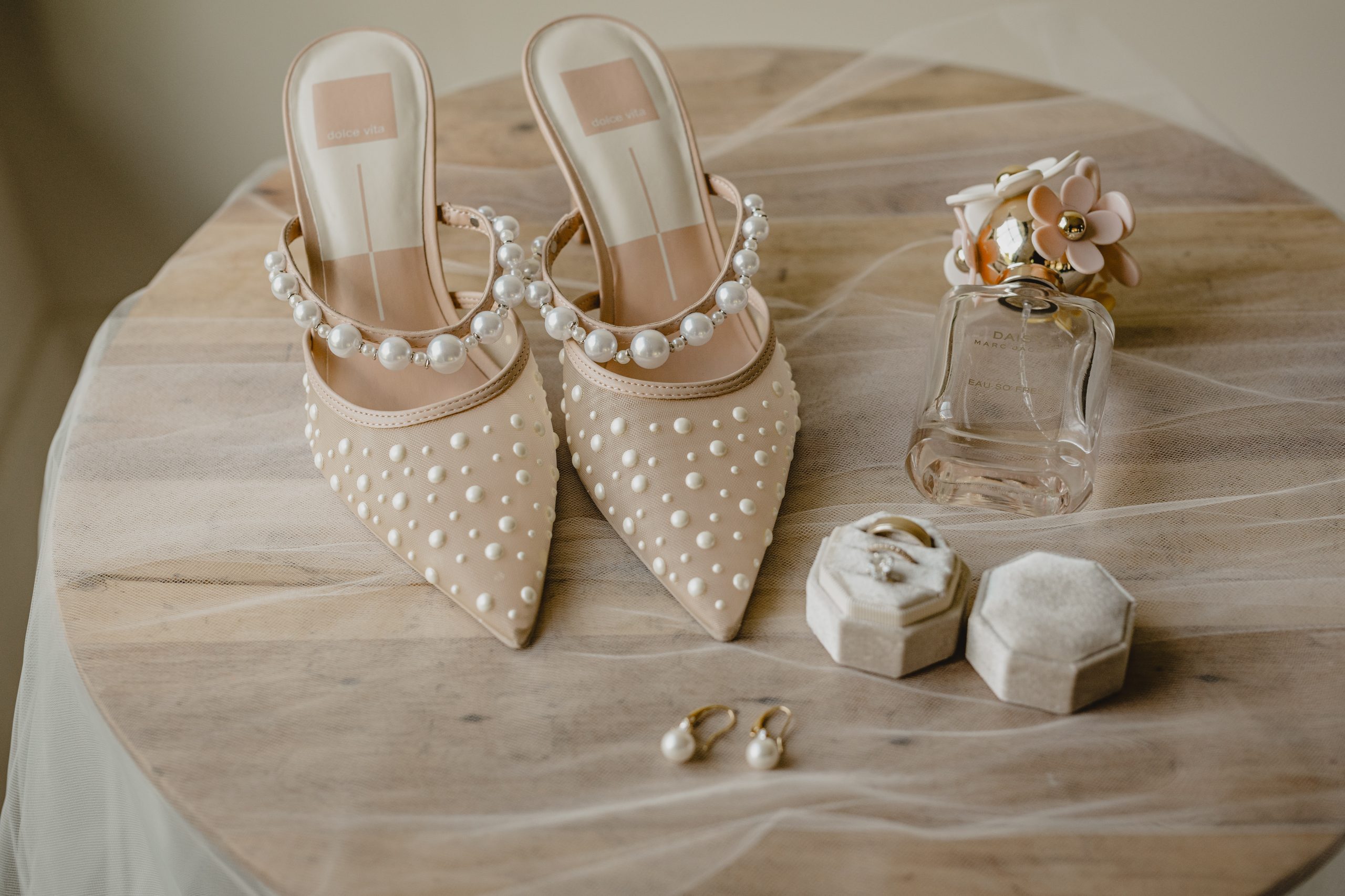 bride's champagne shoes with pearl accents and perfume rest on wooden table with veil underneath 