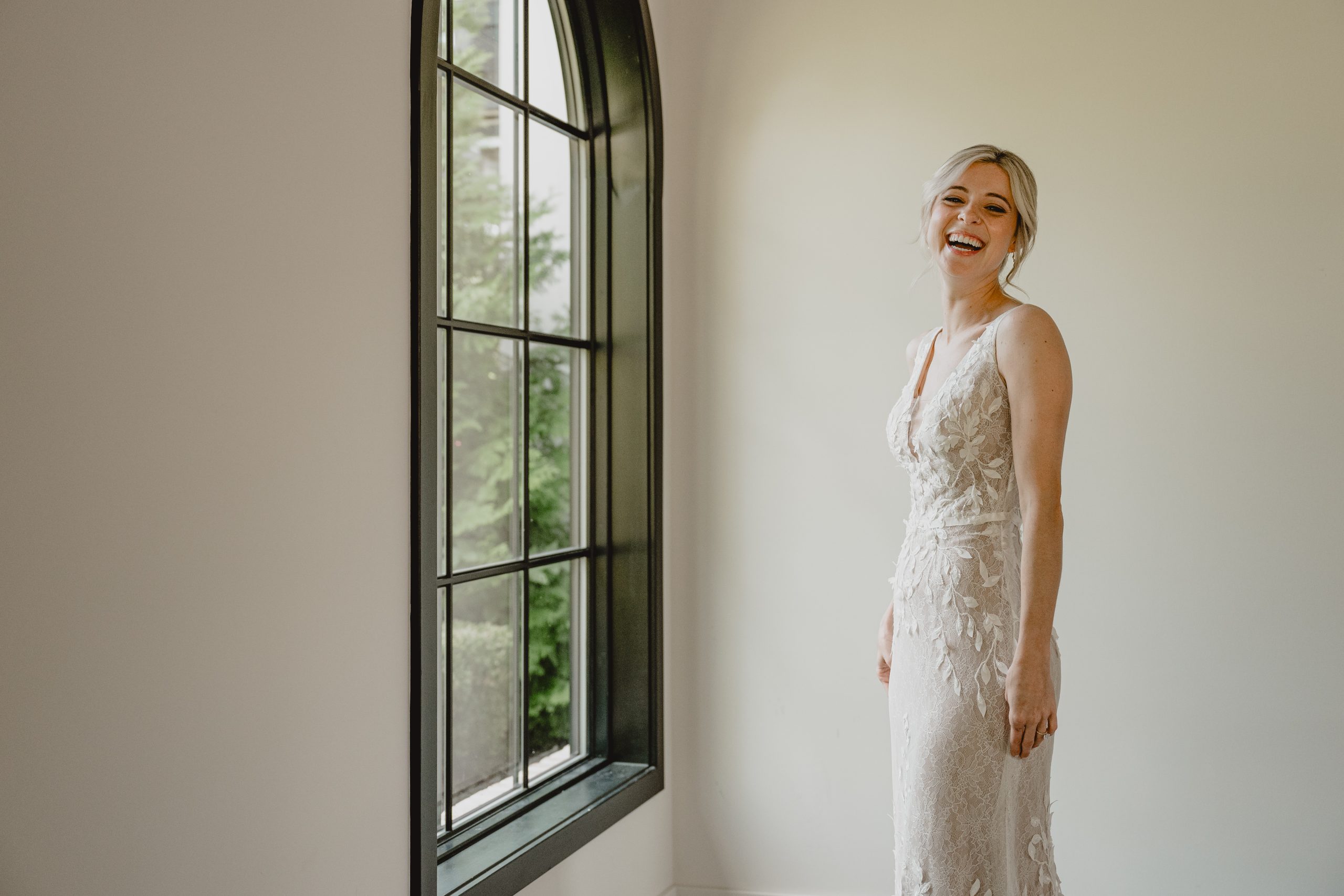 blonde bride in wedding gown with leaves lace overlay laughs in front of window at the Perona Farms Refinery