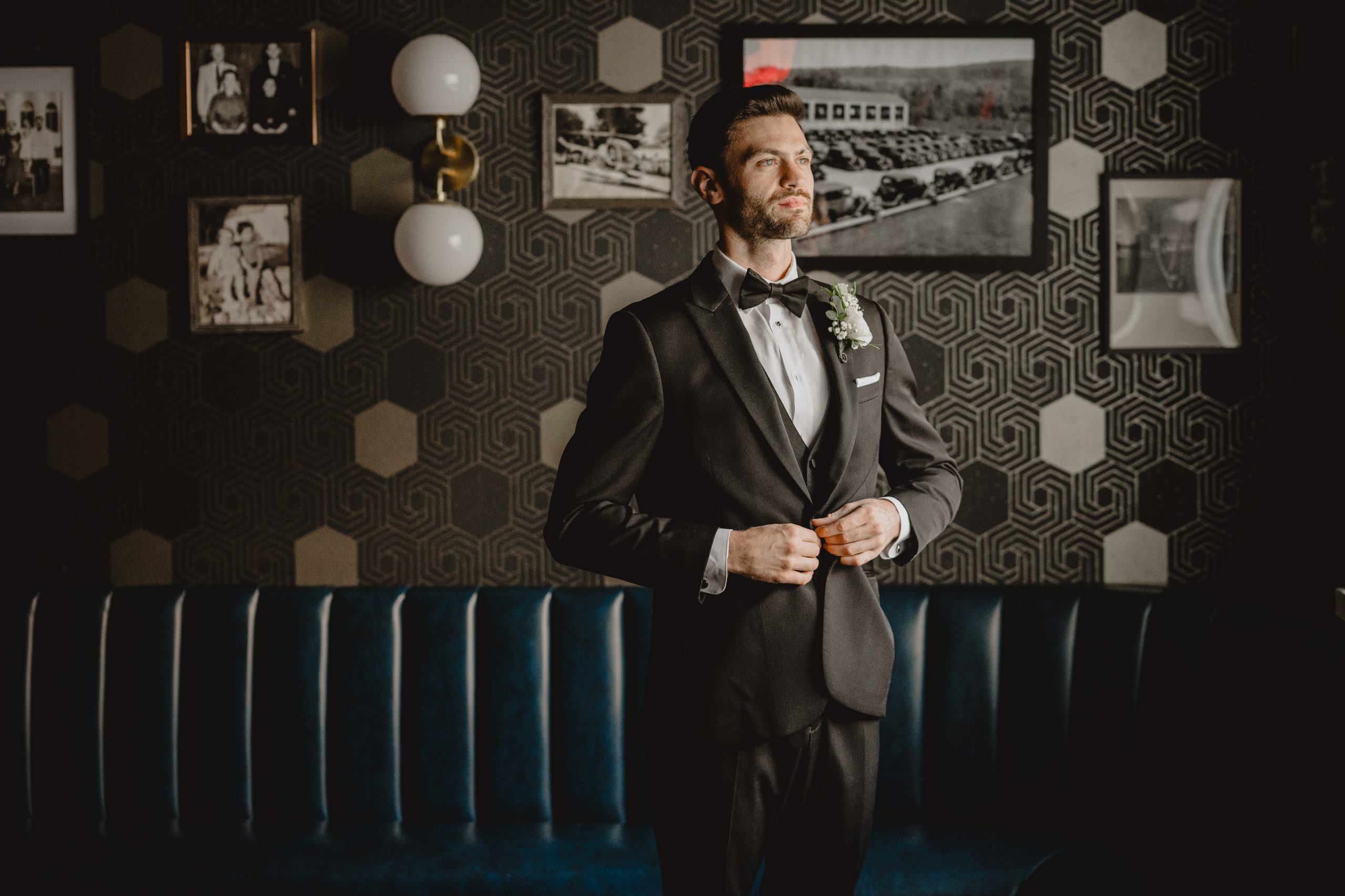 groom buttons up jacket standing in front of black and white wall with blue leather couch