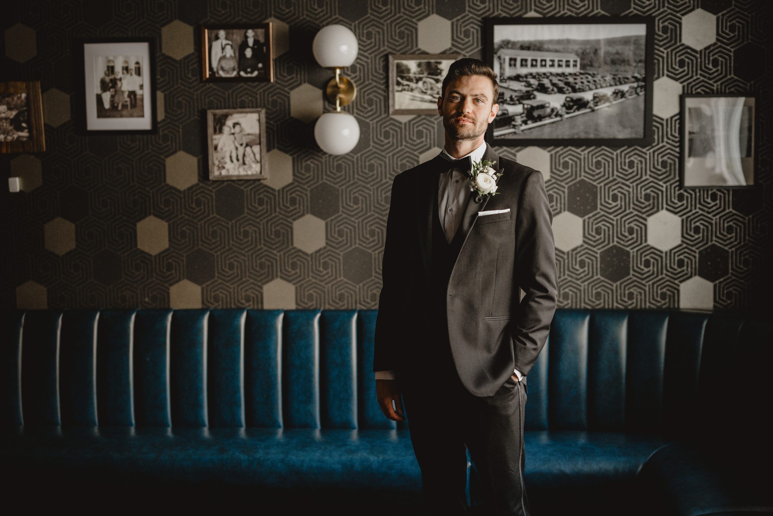 groom stands with hand in pocket of tux jacket in front of tan and black wall with blue couch at the Perona Farms Refinery