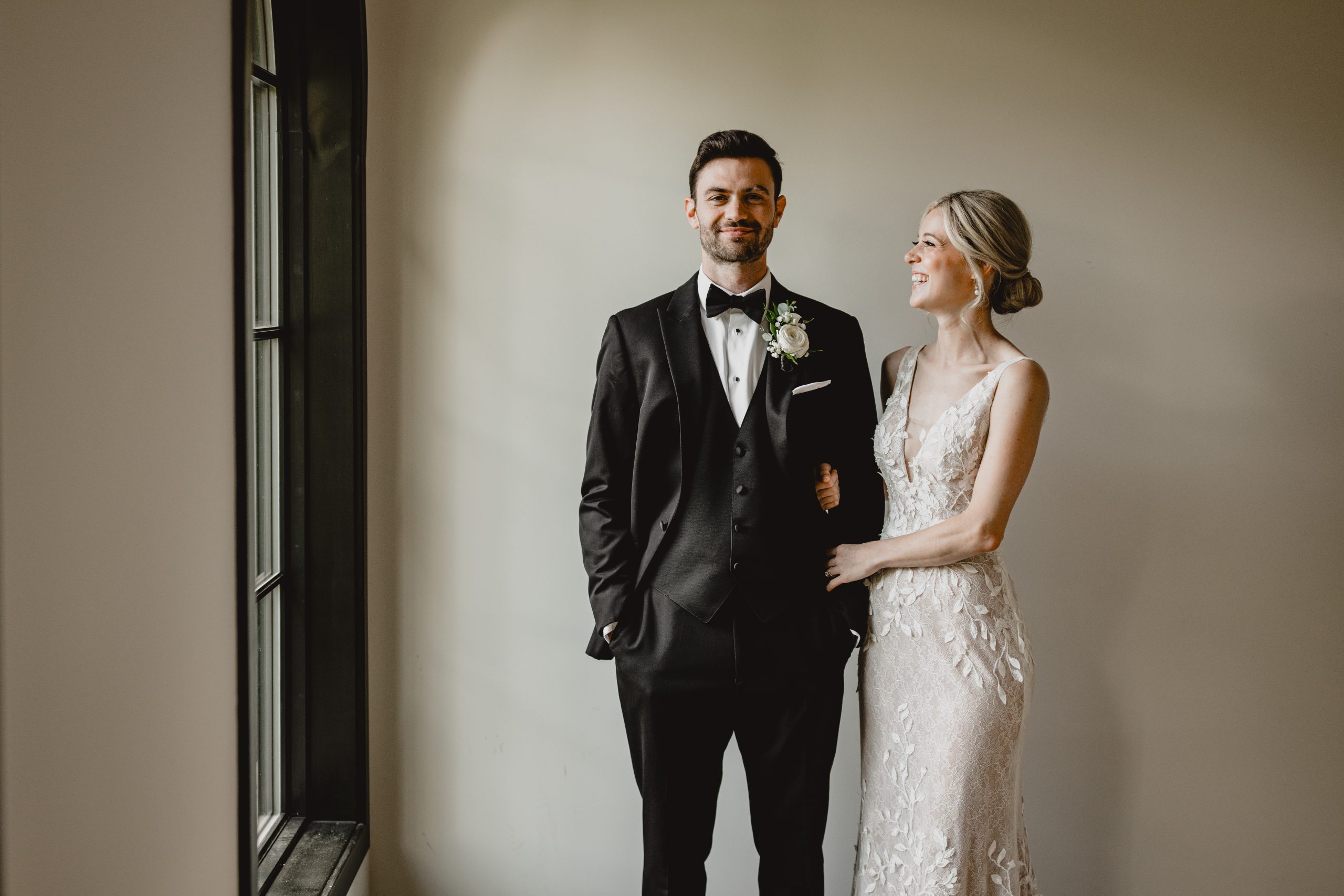 bride holds groom's arm looking at him and laughing during wedding photos by window at the Perona Farms Refinery