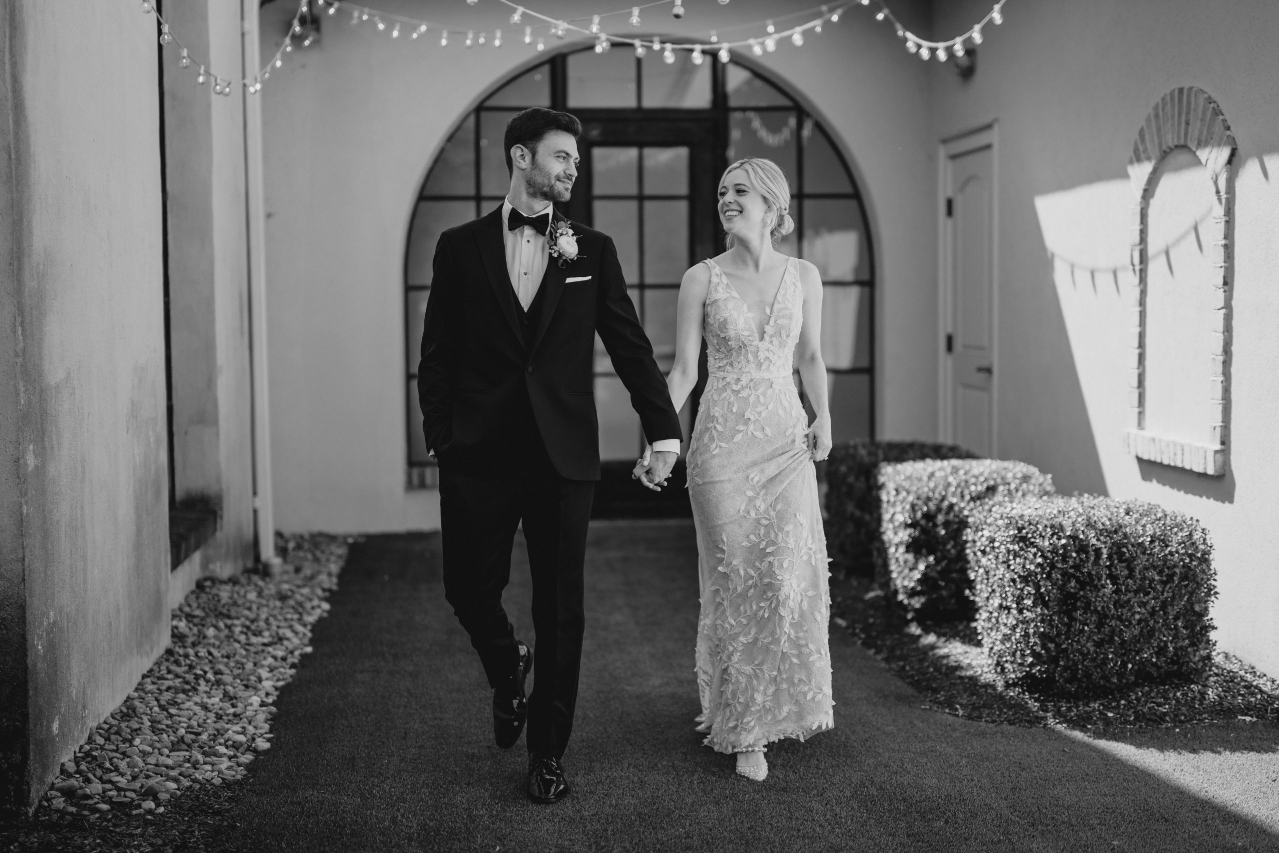 bride and groom hold hands walking in courtyard with bistro lights above them 