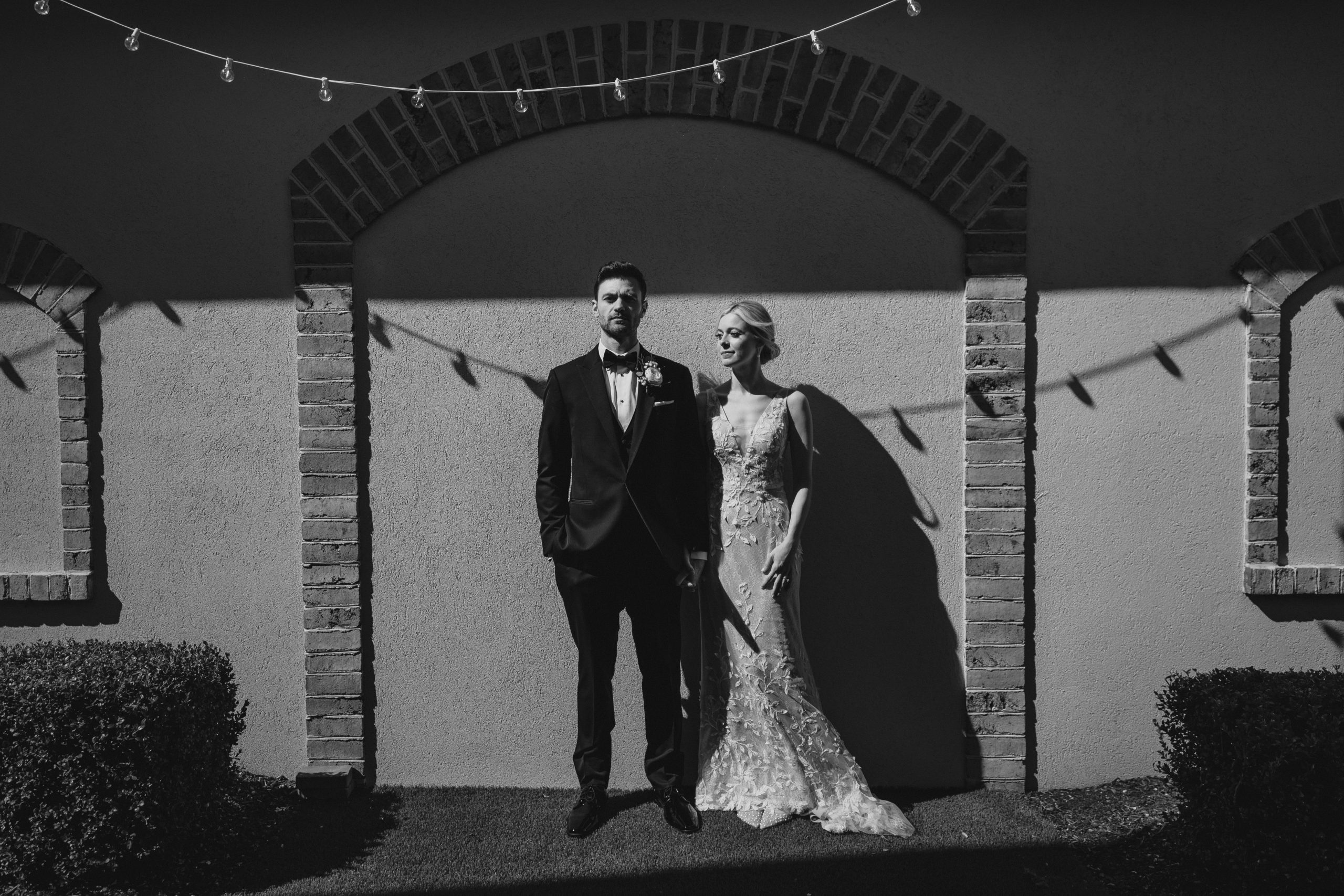 newlyweds stand in front of wall with shadow of bistro lights over them at the Perona Farms Refinery