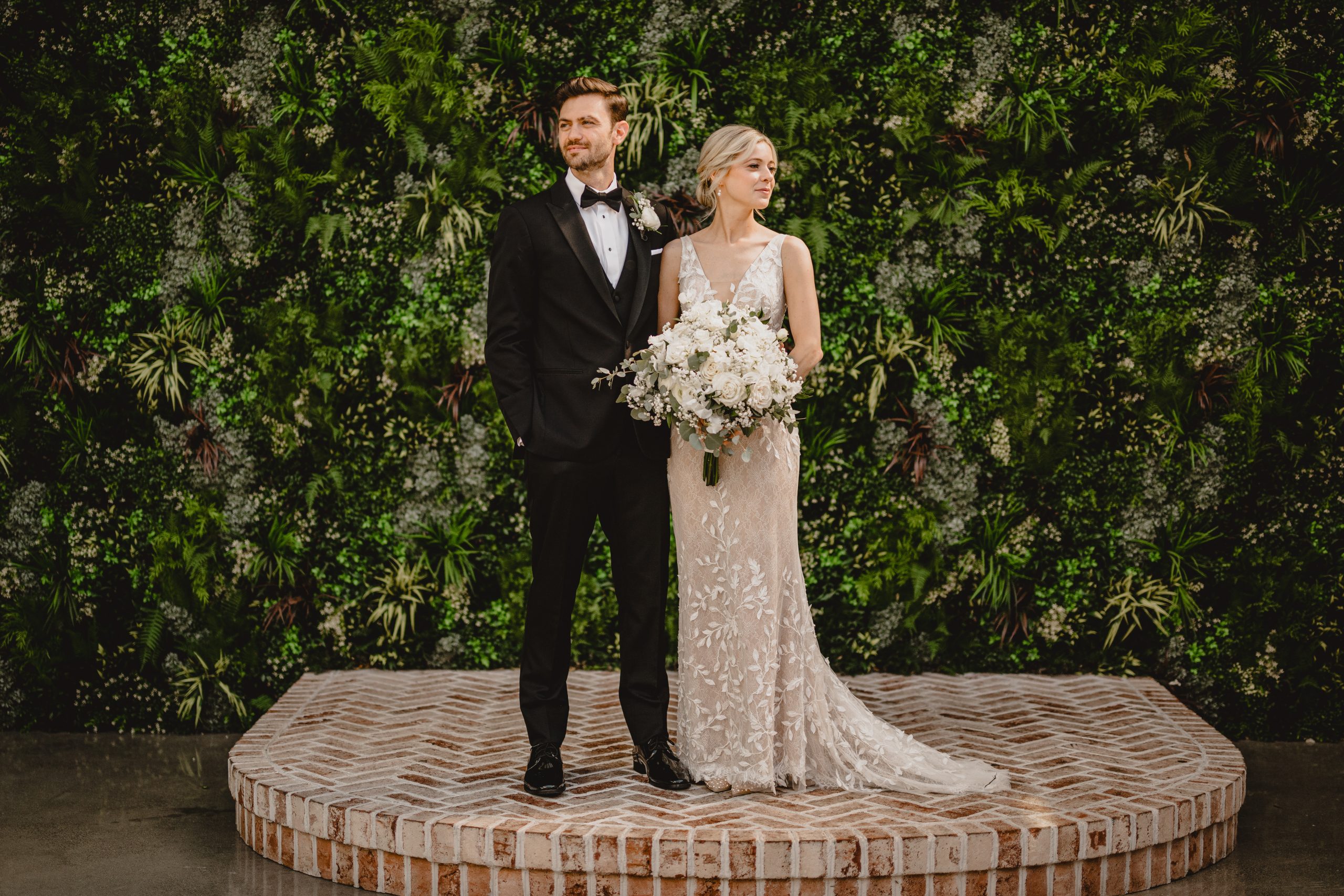 newlyweds stand on brick dais in courtyard of the Perona Farms Refinery looking away from each other 