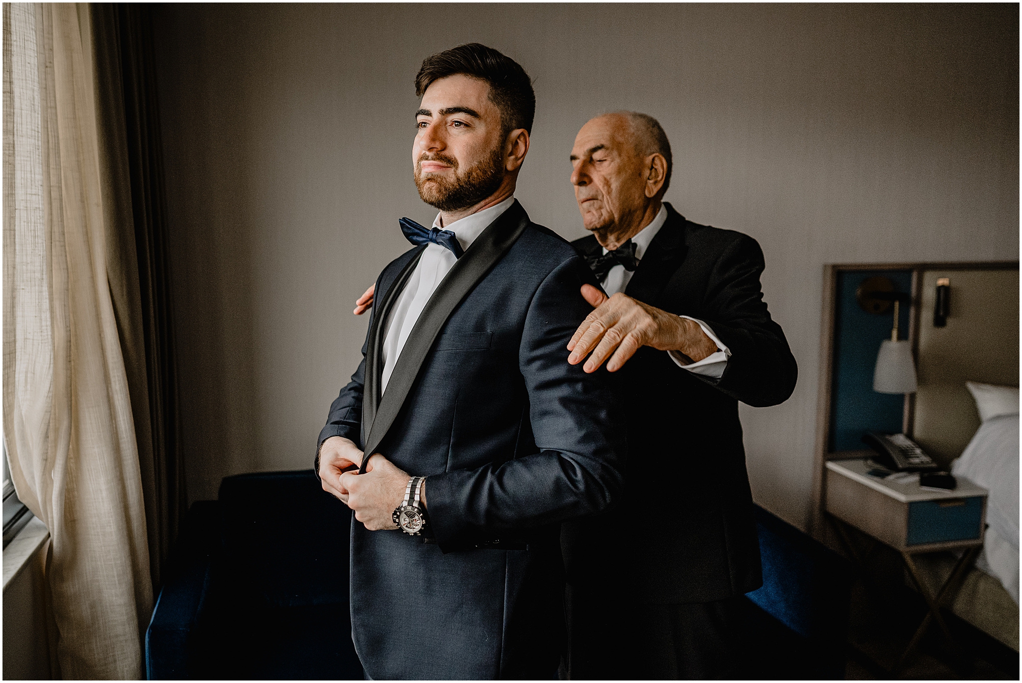 father helps groom into navy blue tux jacket during prep for NYC wedding day