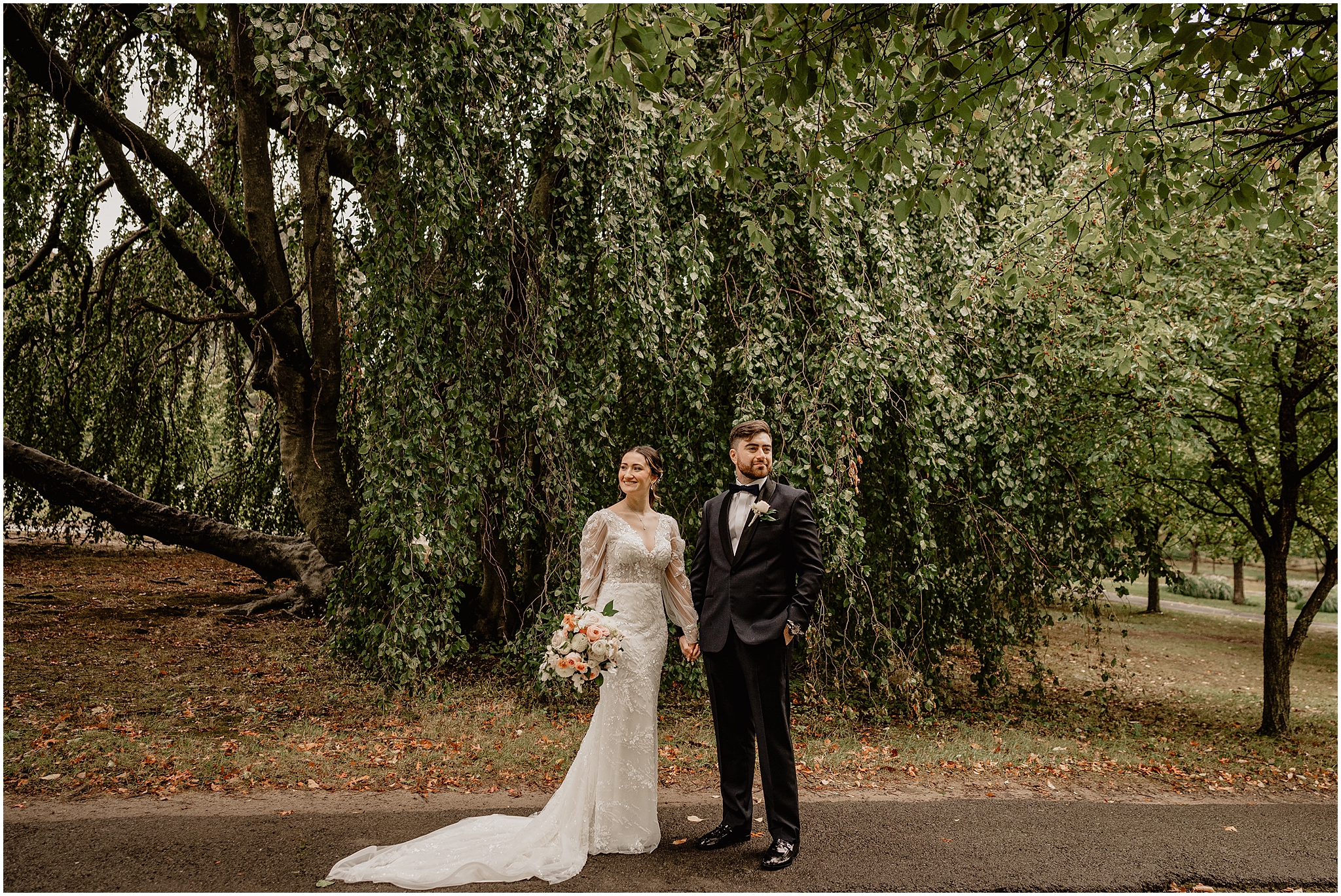 bride and groom hold hands looking away from each other in front of weeping willow tree