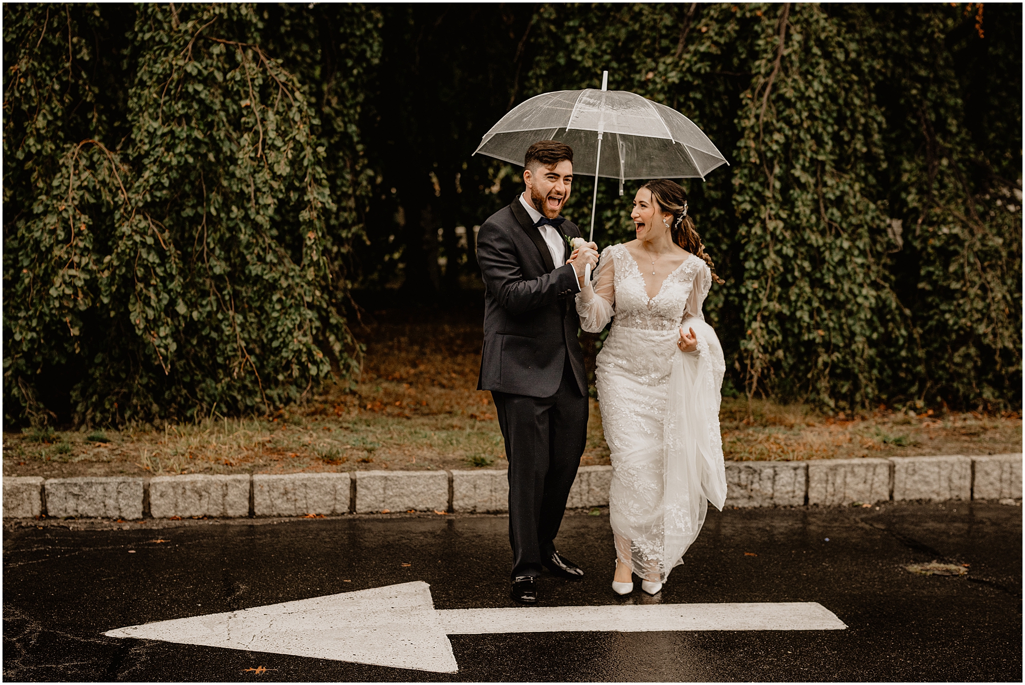 newlyweds laugh walking on street under clear umbrella on rainy wedding day at Whitby Castle