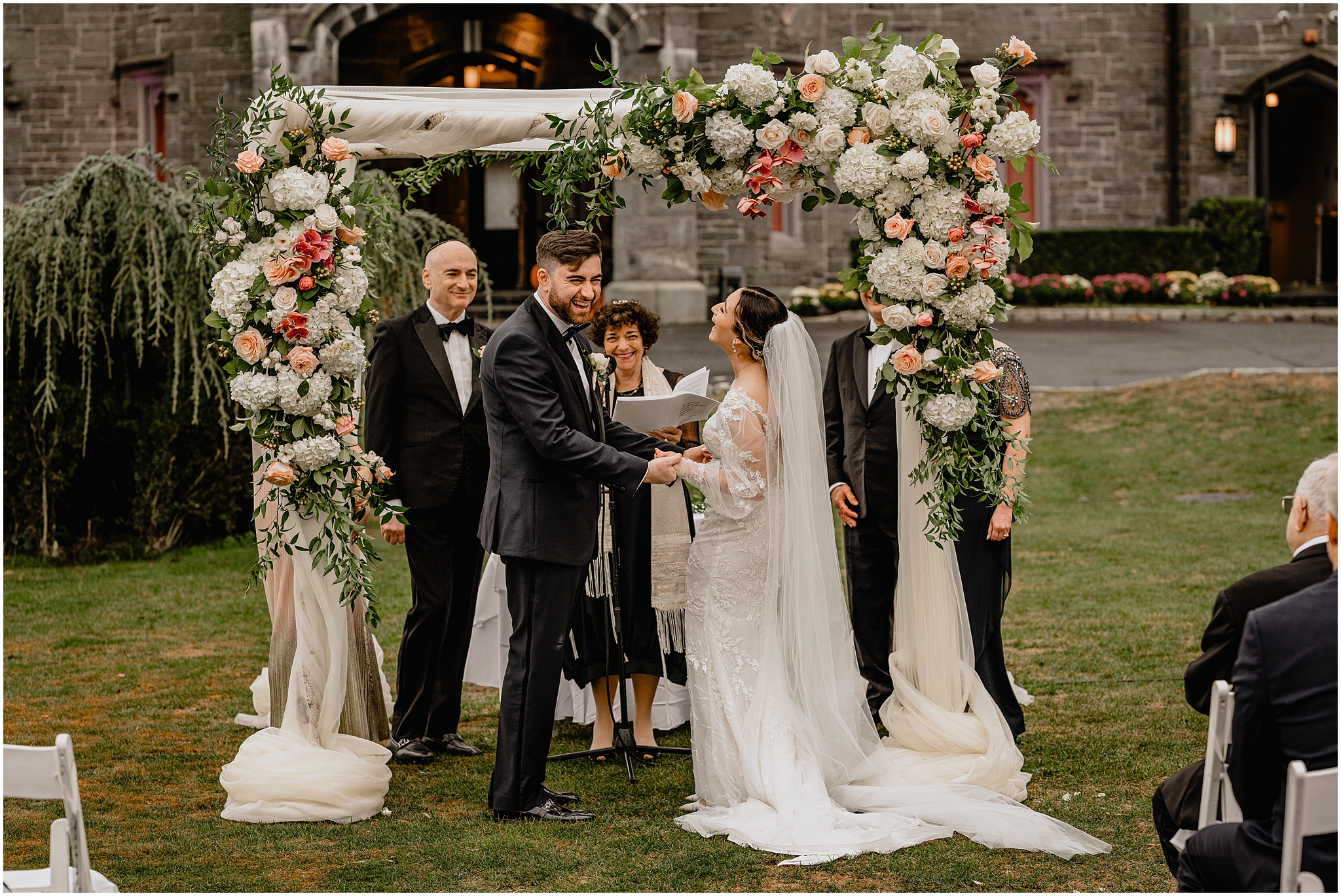 newlyweds hold hands laughing under chuppah during Jewish wedding ceremony on the lawn at Whitby Castle