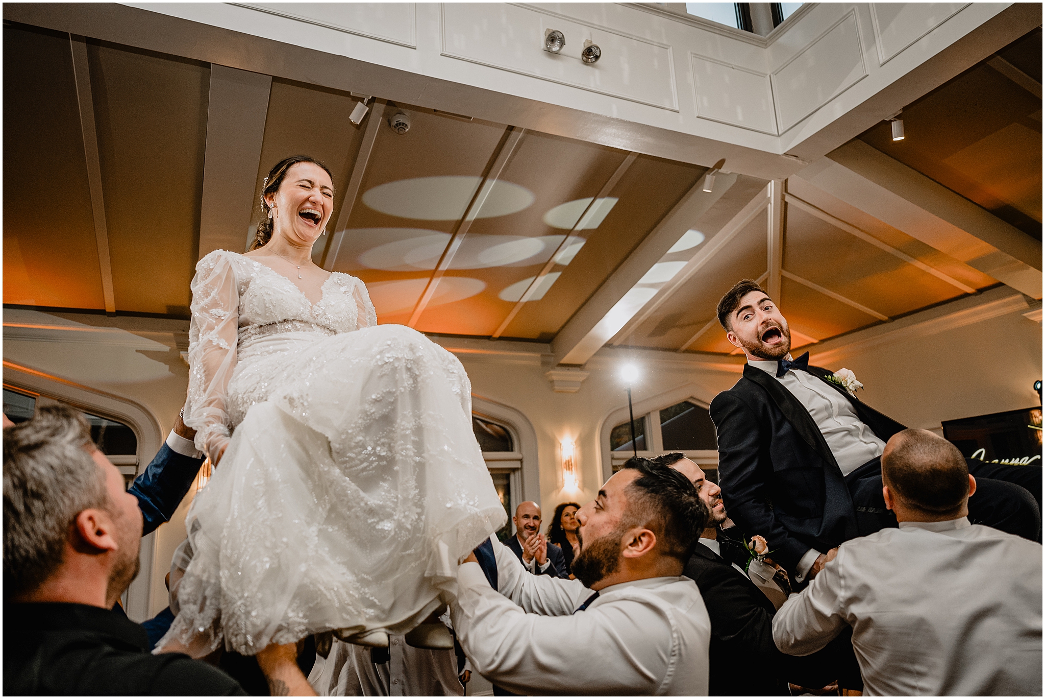wedding guests lift up bride and groom in chairs during hora 