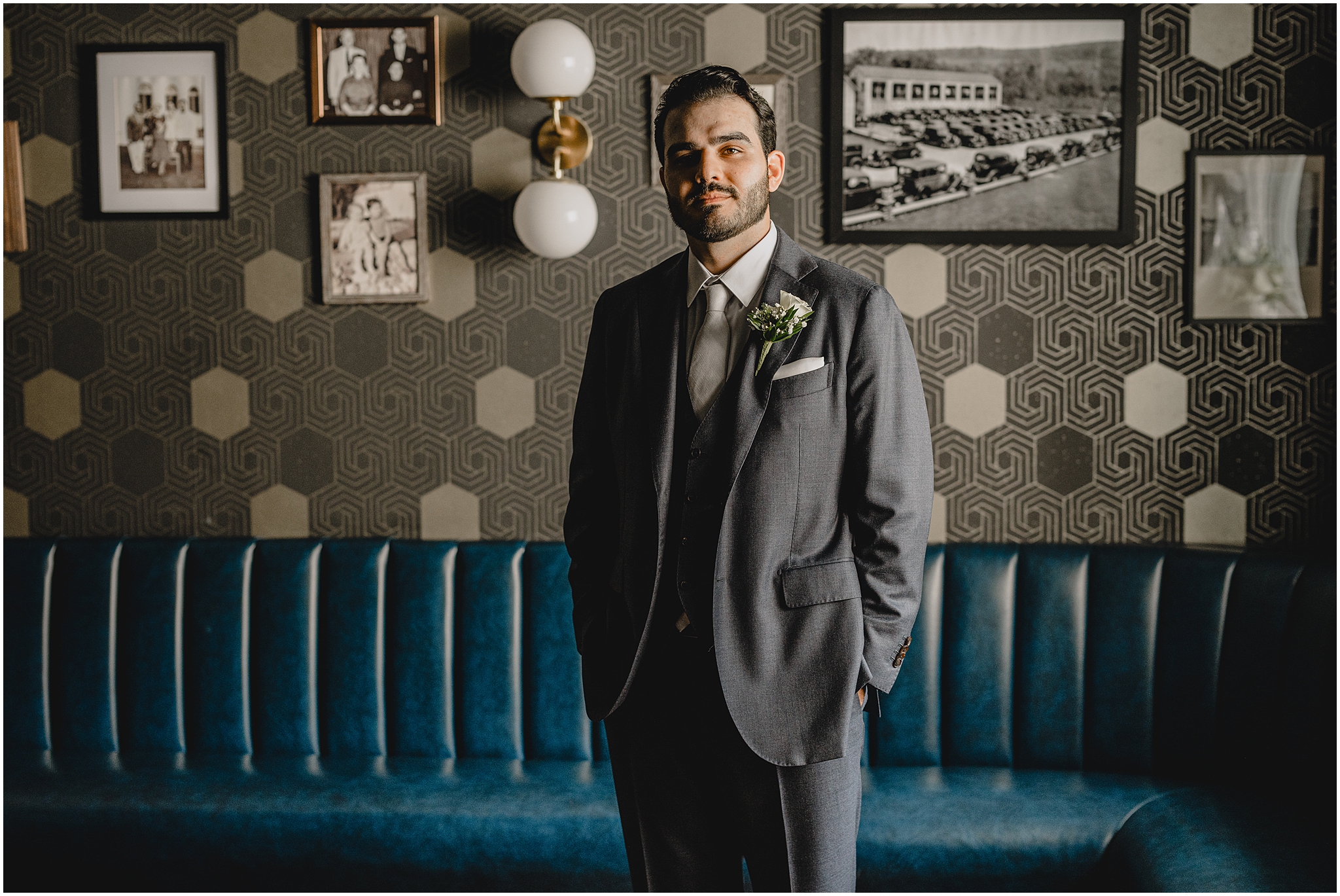 groom stands with hands in pockets of suit in front of blue leather couch and geometric wall at the Refinery at Perona Farms