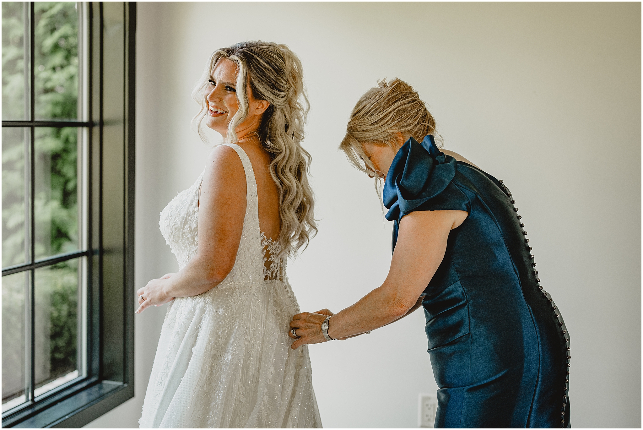 mother in navy blue dress helps bride button up the back of her wedding dress