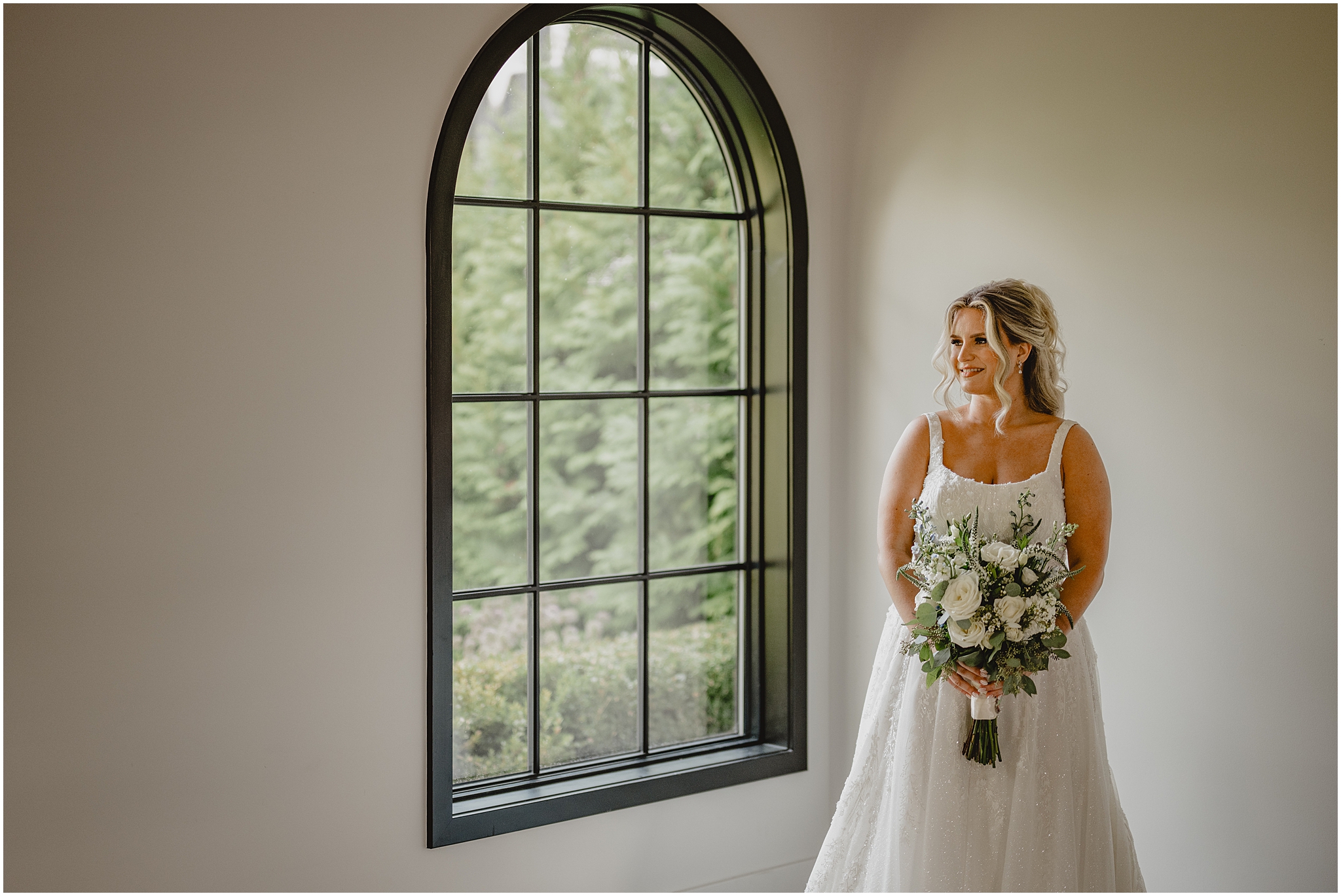 bride stands by grey window at the Refinery at Perona Farms holding bouquet of white roses and greenery 